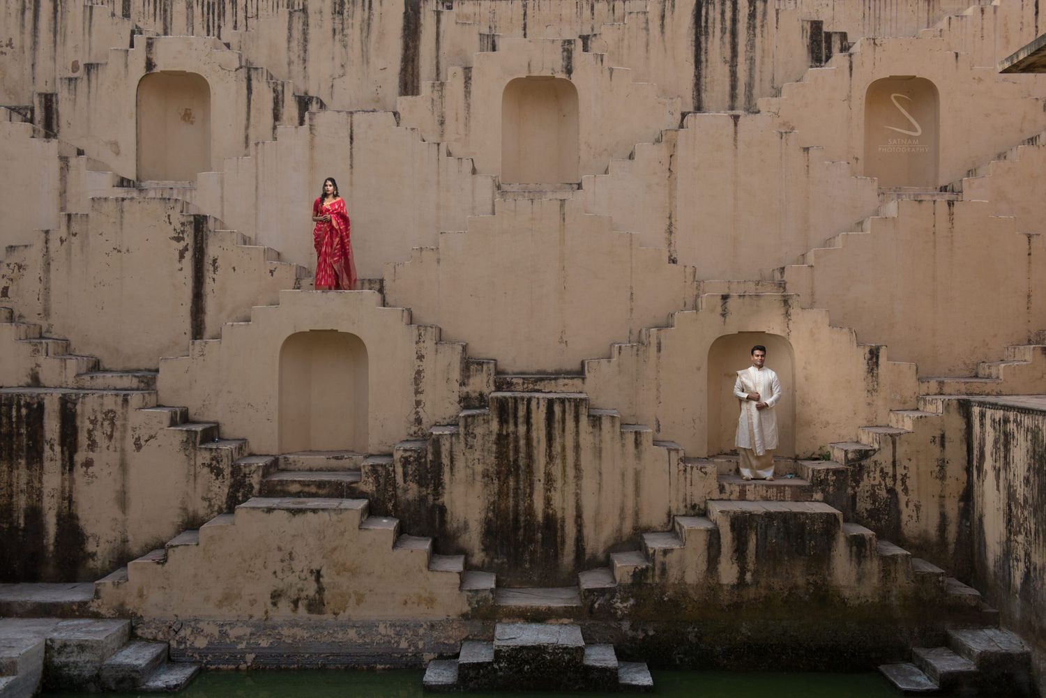 Couple in traditional Indian red and white outfits posing separately on the steps of an ancient stepwell.