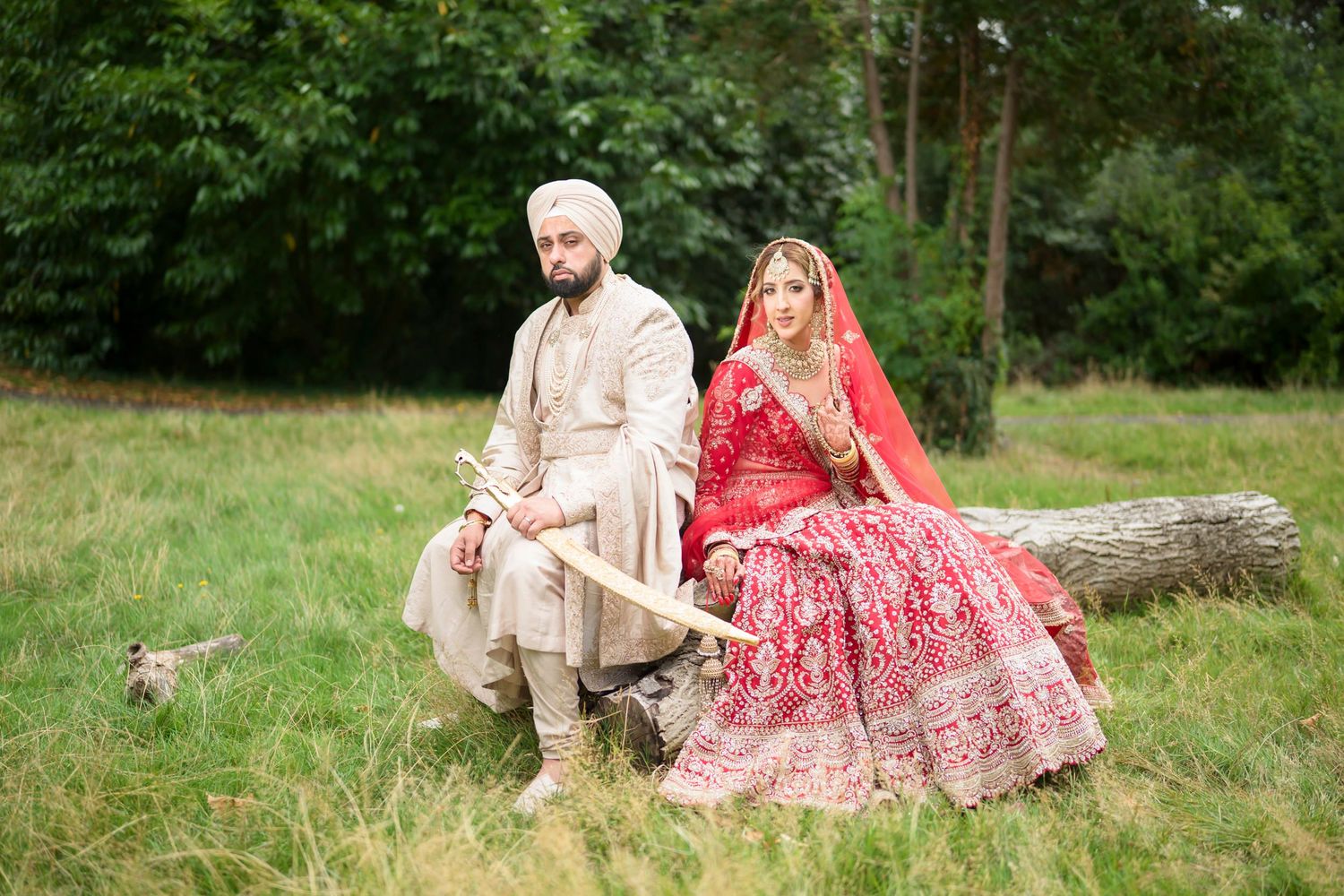Sikh bride in red lehenga and groom in cream sherwani sit on log outdoors.