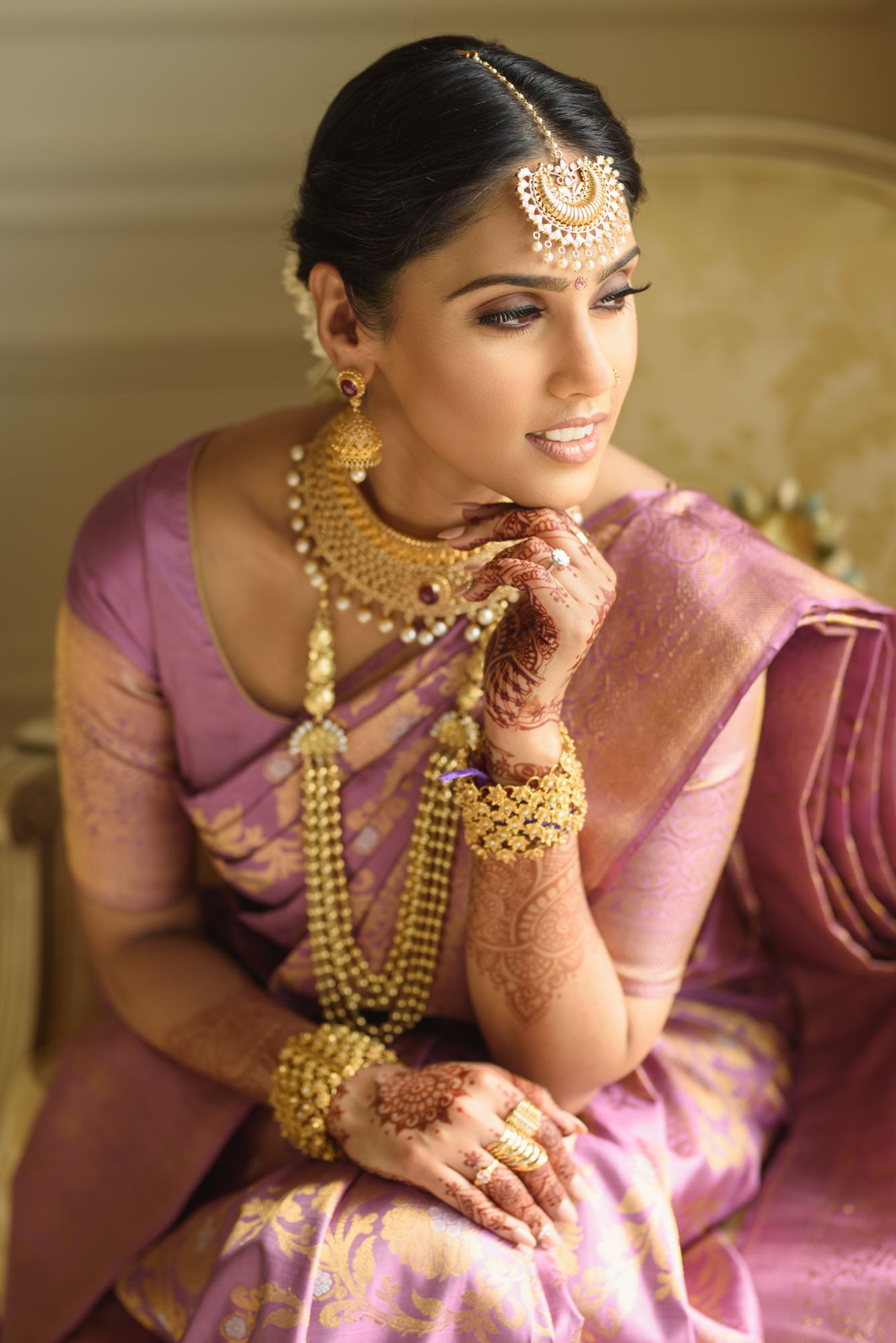 South Indian bride in purple Kanjivaram saree with gold jewelry posing in elegant seated pose.