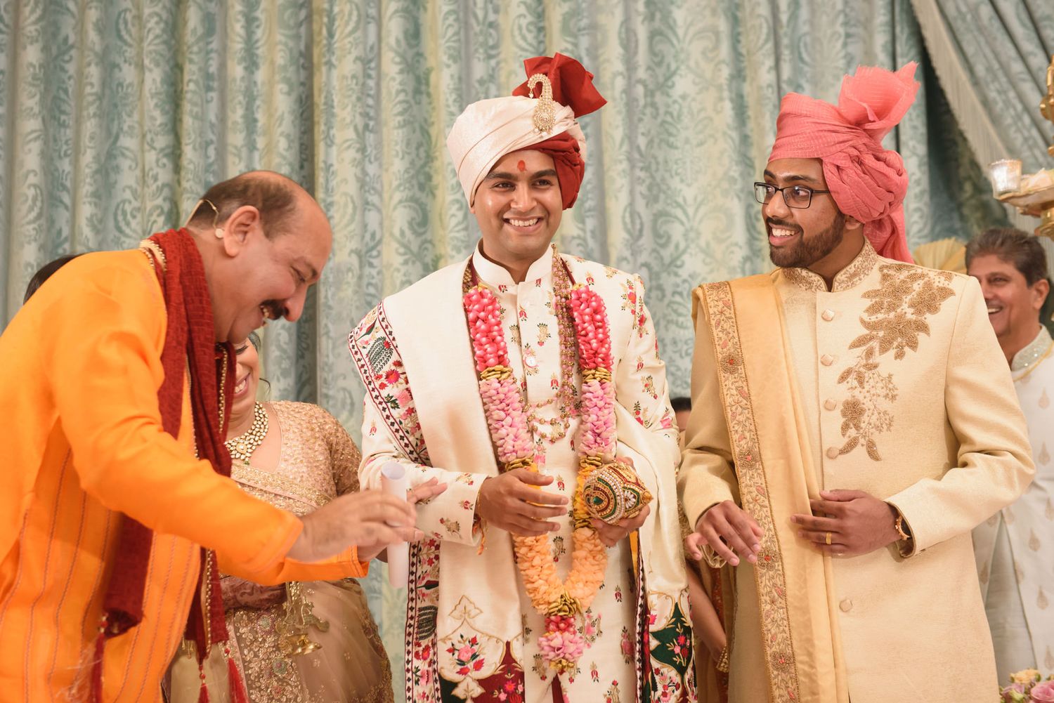 Indian groom in floral sherwani smiles during traditional Hindu wedding ceremony at ornate venue.
