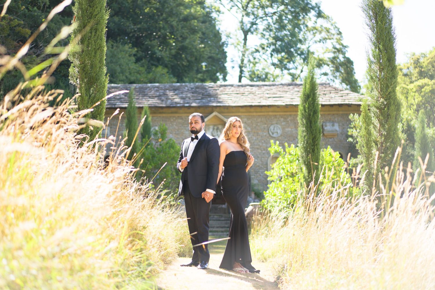 Elegant couple in black formal attire walking through lush garden with cypress trees at golden hour.