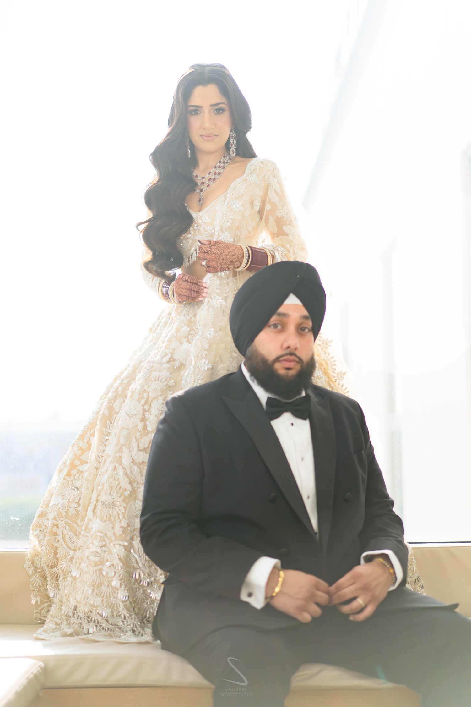 A Sikh bride in an ivory embroidered lehenga stands behind her groom in a black tuxedo by a bright window.