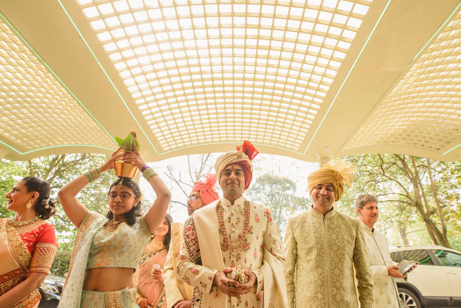 Indian groom in floral sherwani walks under decorative canopy during traditional wedding procession.