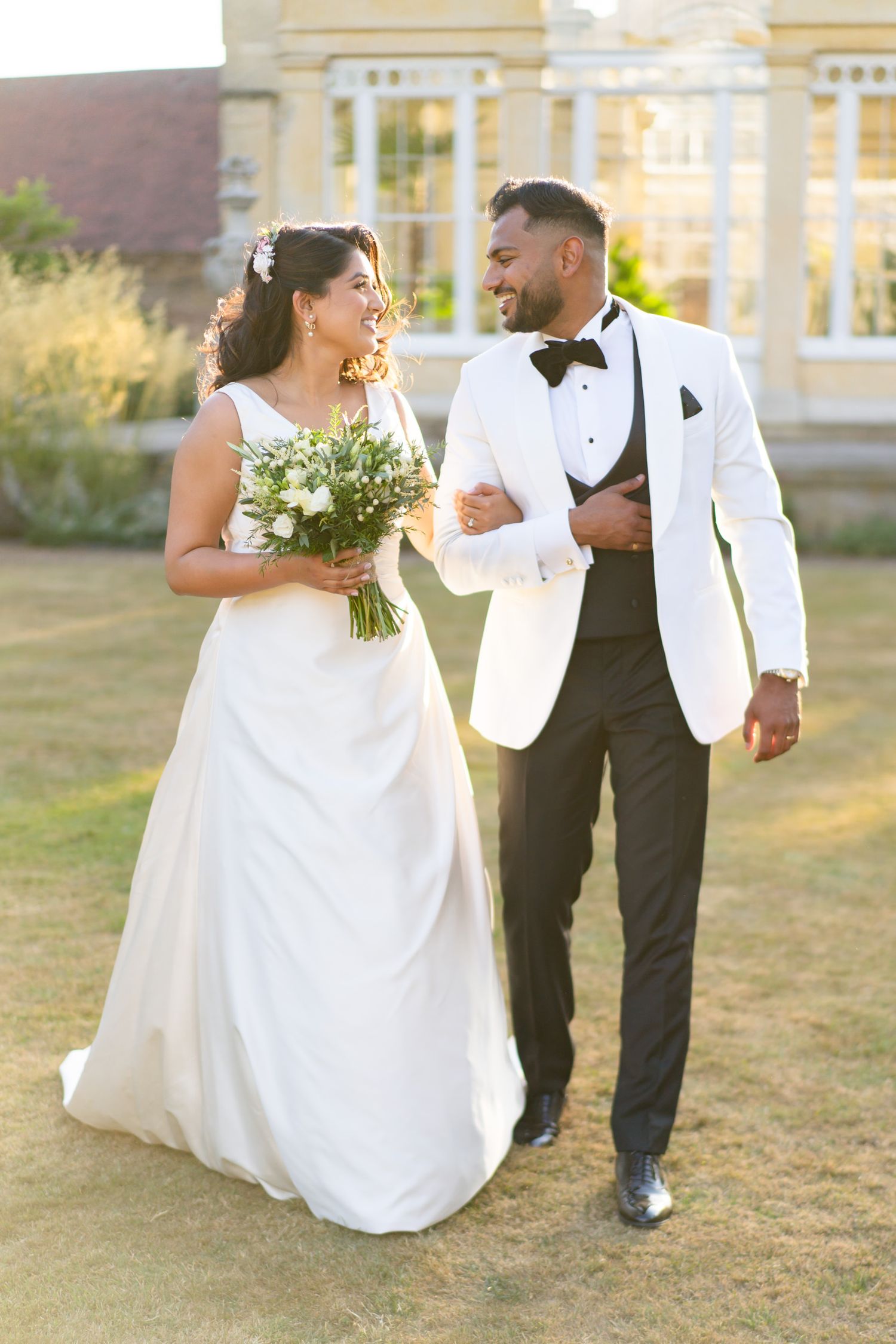 A bride in a white gown holding a bouquet walks arm in arm with her groom in a white tuxedo jacket outdoors.
