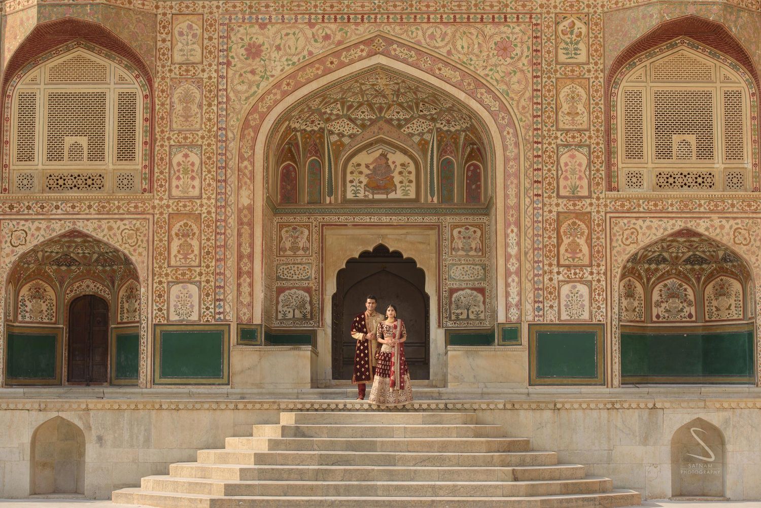 Indian couple in wedding attire poses on steps before the intricately decorated facade of Amer Fort.