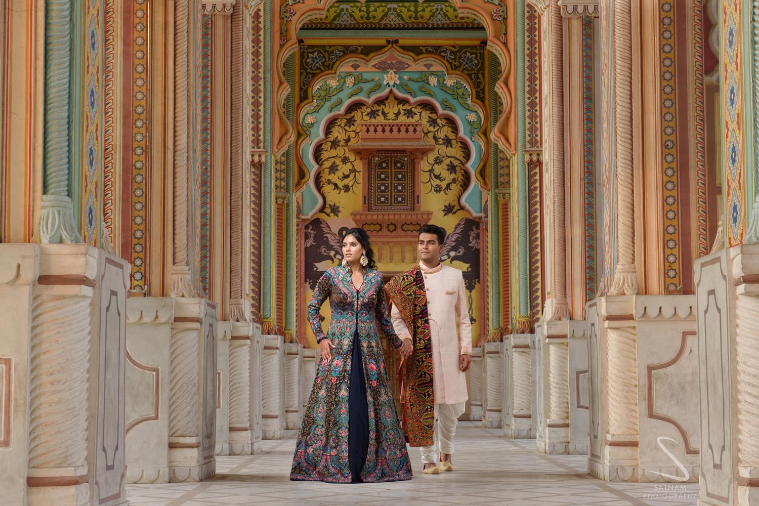 Couple in royal Indian traditional attire walking through vibrant painted palace corridor.