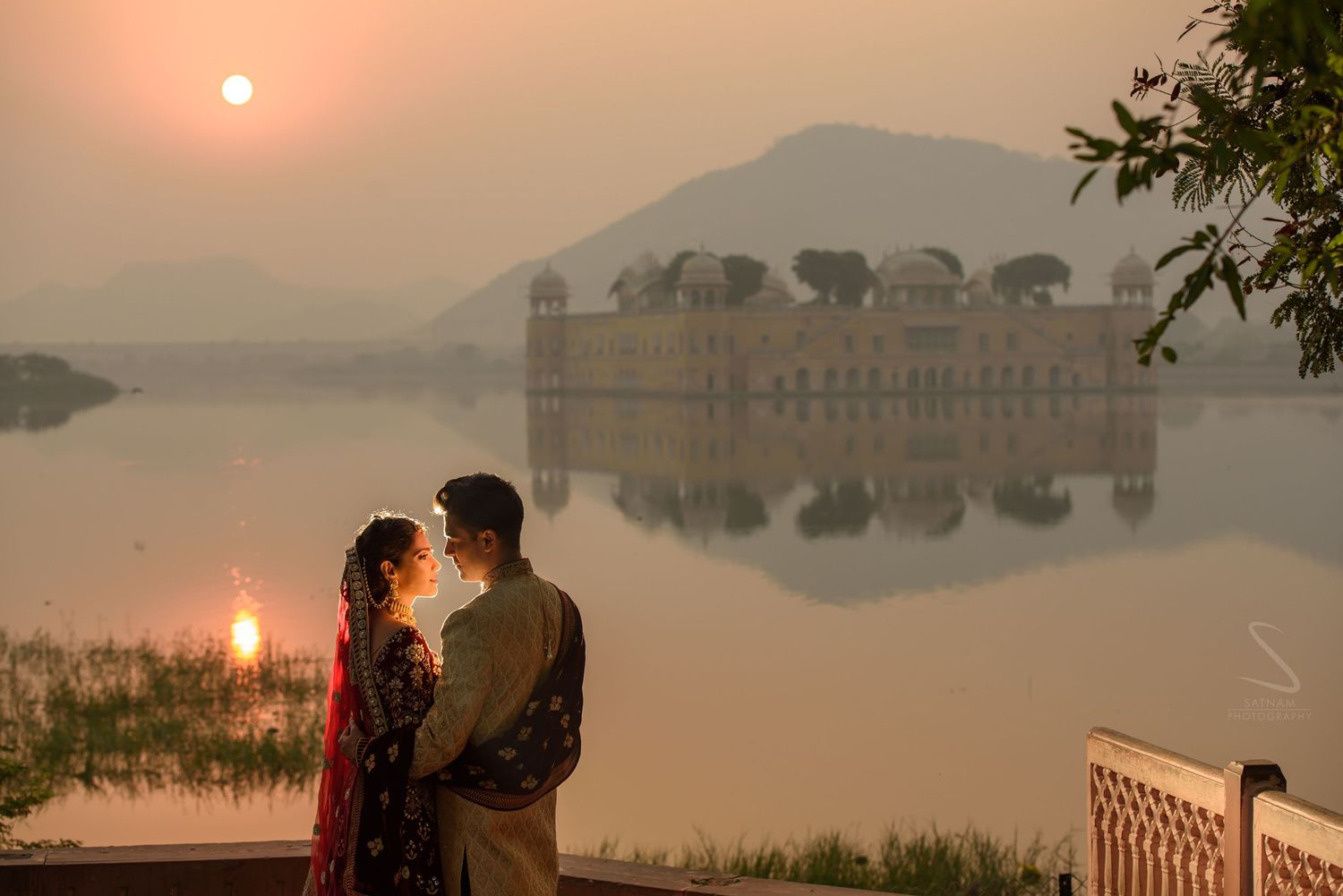 Indian couple in traditional wedding attire embraces at sunset near Jal Mahal palace in Jaipur.