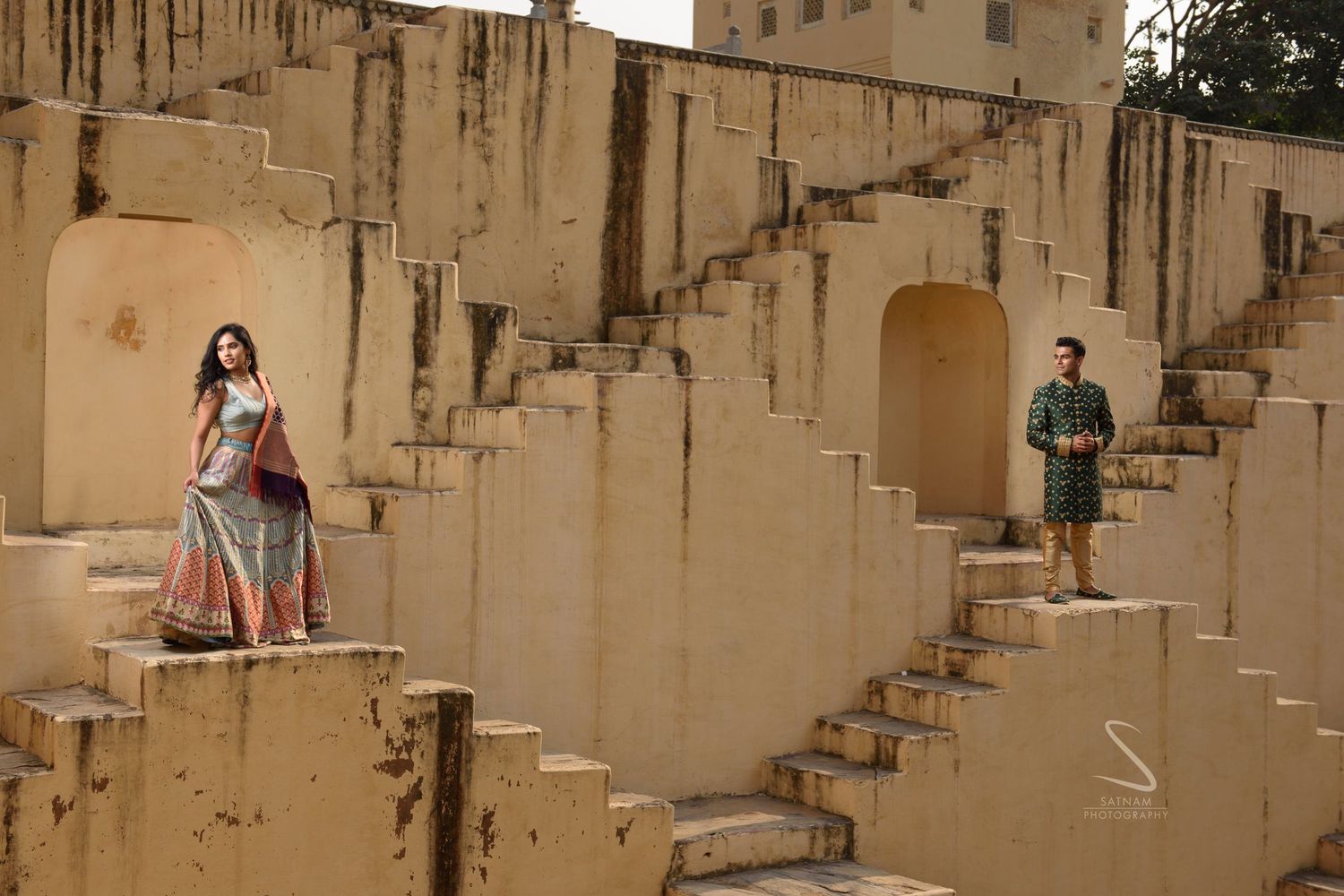 Couple in traditional Indian outfits posing on ancient stepwell stone architecture.