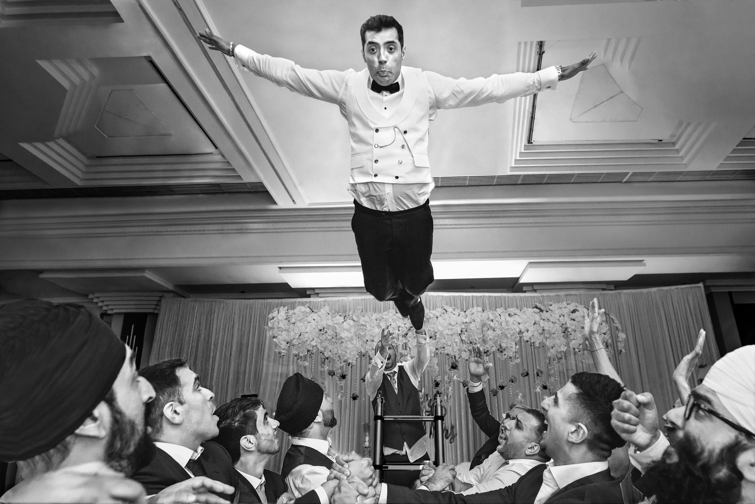 Groom tossed in air by groomsmen during lively black and white wedding reception.