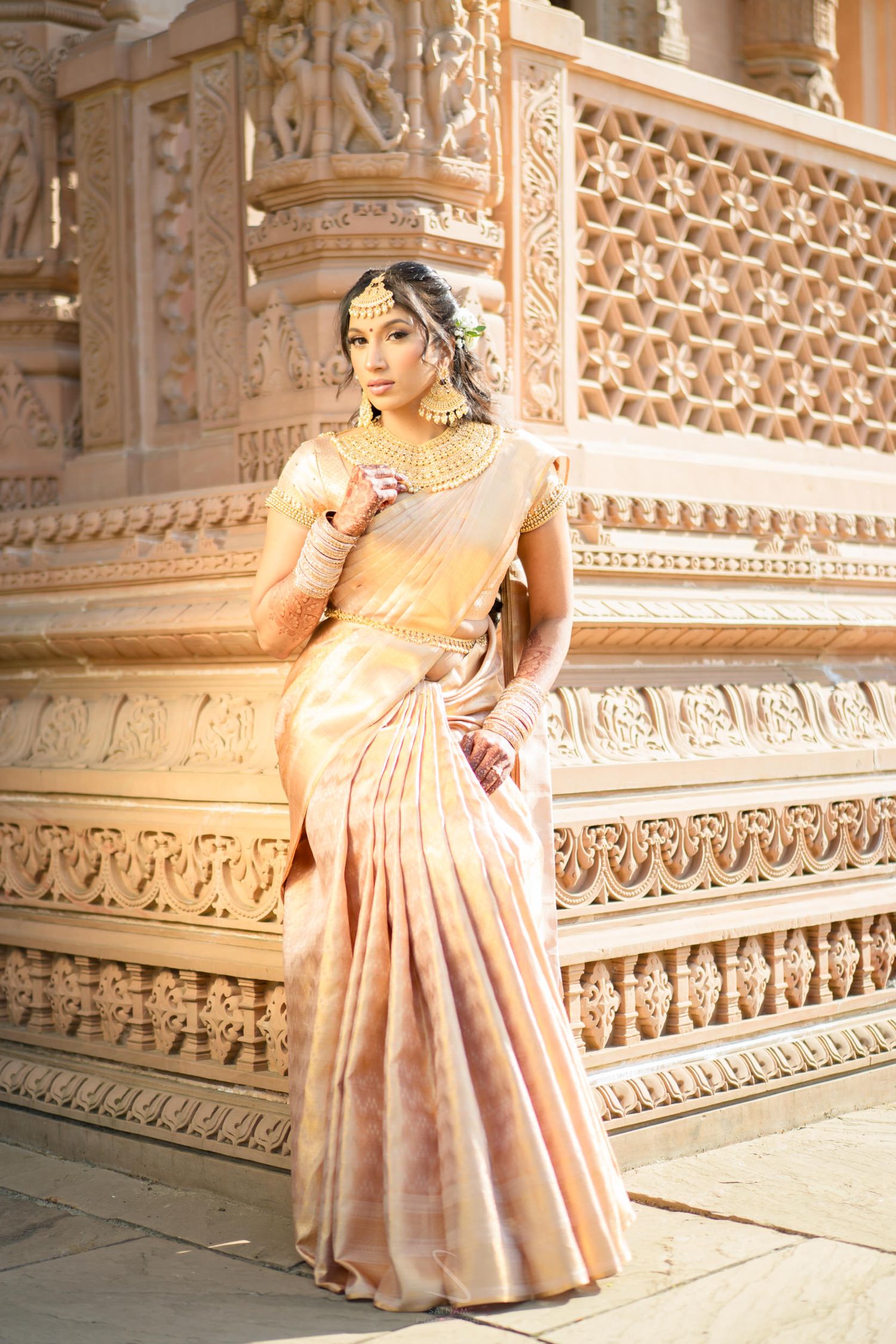 Elegant bride in golden silk saree seated on ornate temple steps with intricate stone carvings.