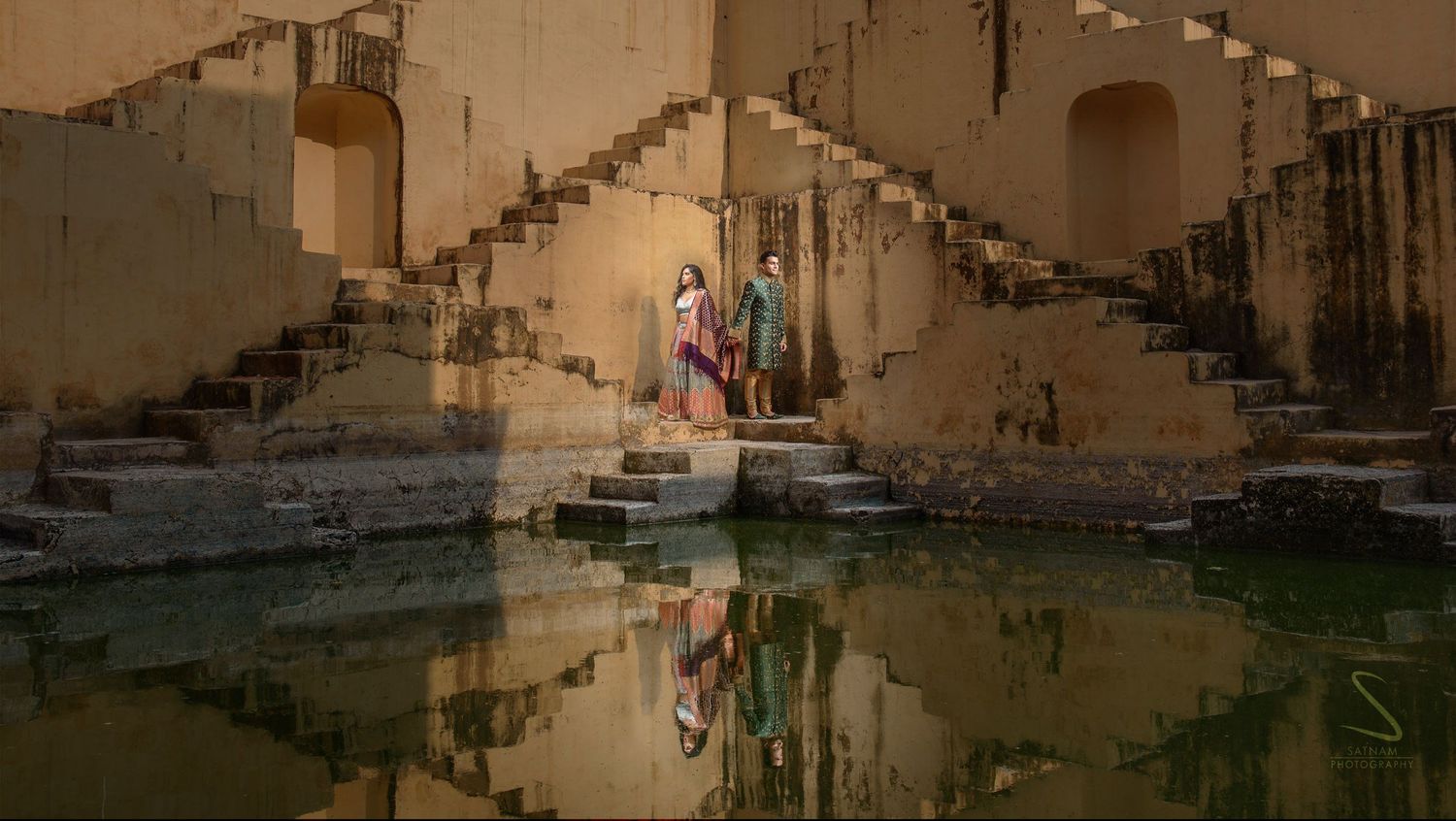 Couple in colorful Indian outfits stands by a reflective green pool inside a Jaipur stepwell.