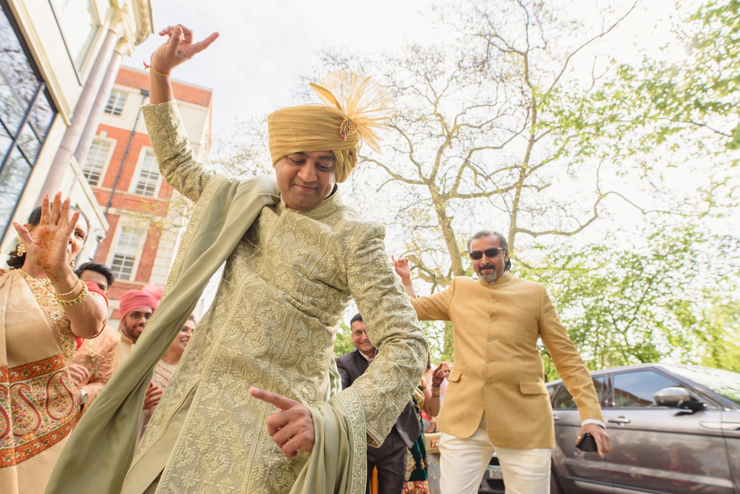 Indian groom in gold sherwani and yellow turban dances joyfully during baraat wedding procession.
