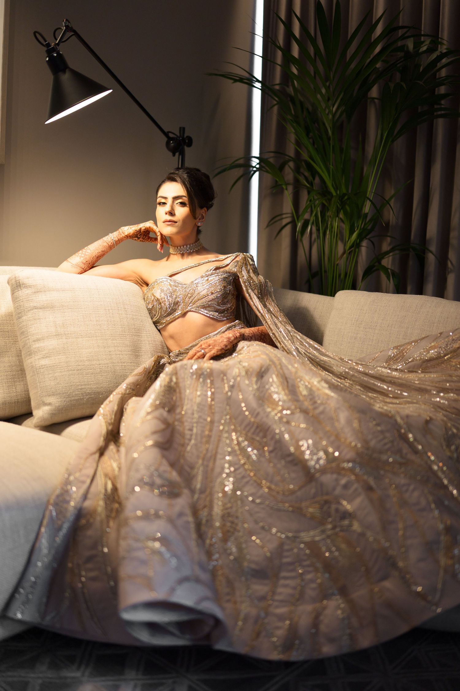 Bride in silver sequined lehenga reclines elegantly on white sofa under dramatic studio lighting.