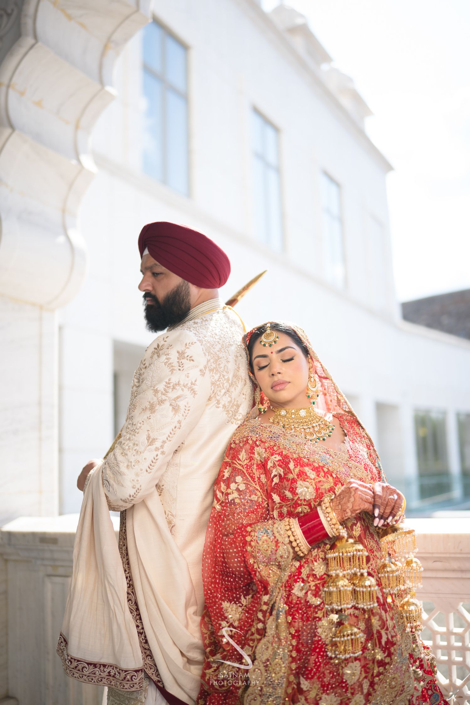 Sikh bride and groom in traditional red and cream wedding attire pose back-to-back at white building.