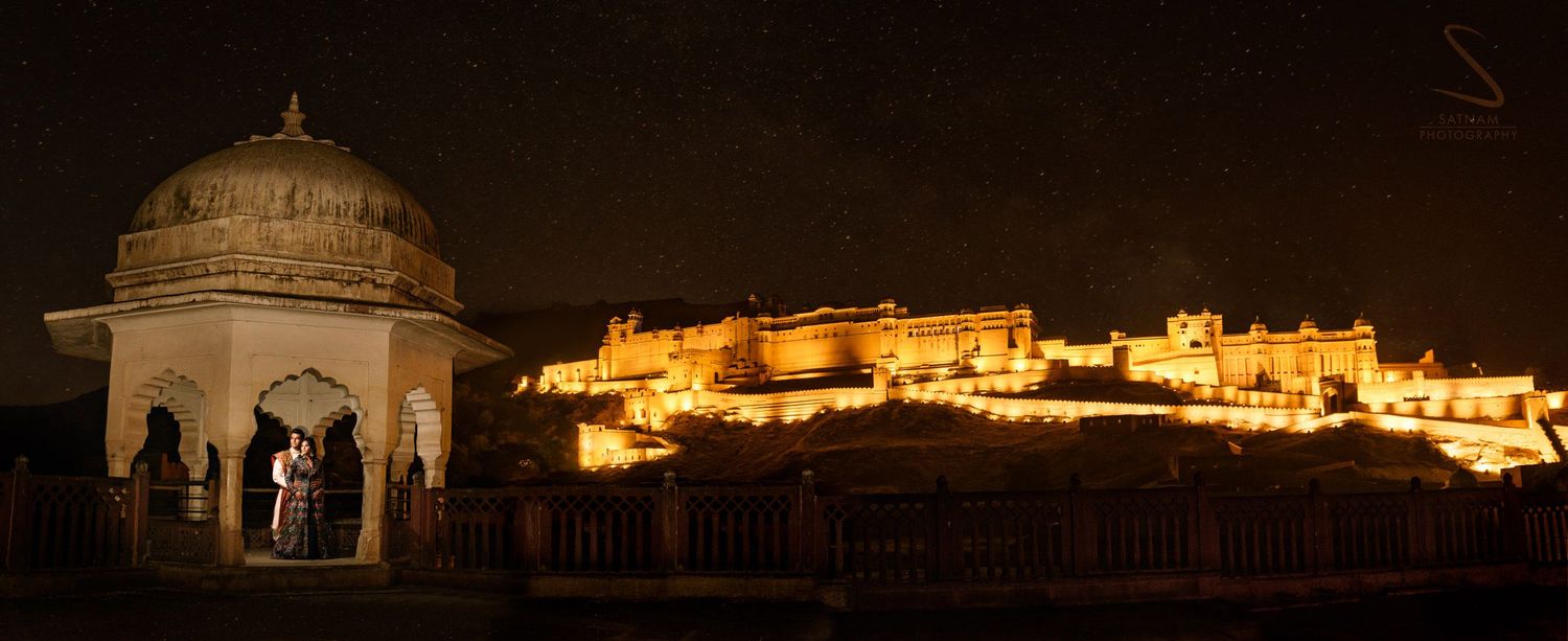 Couple standing before illuminated Amber Fort at night under starry sky in Jaipur, Rajasthan.