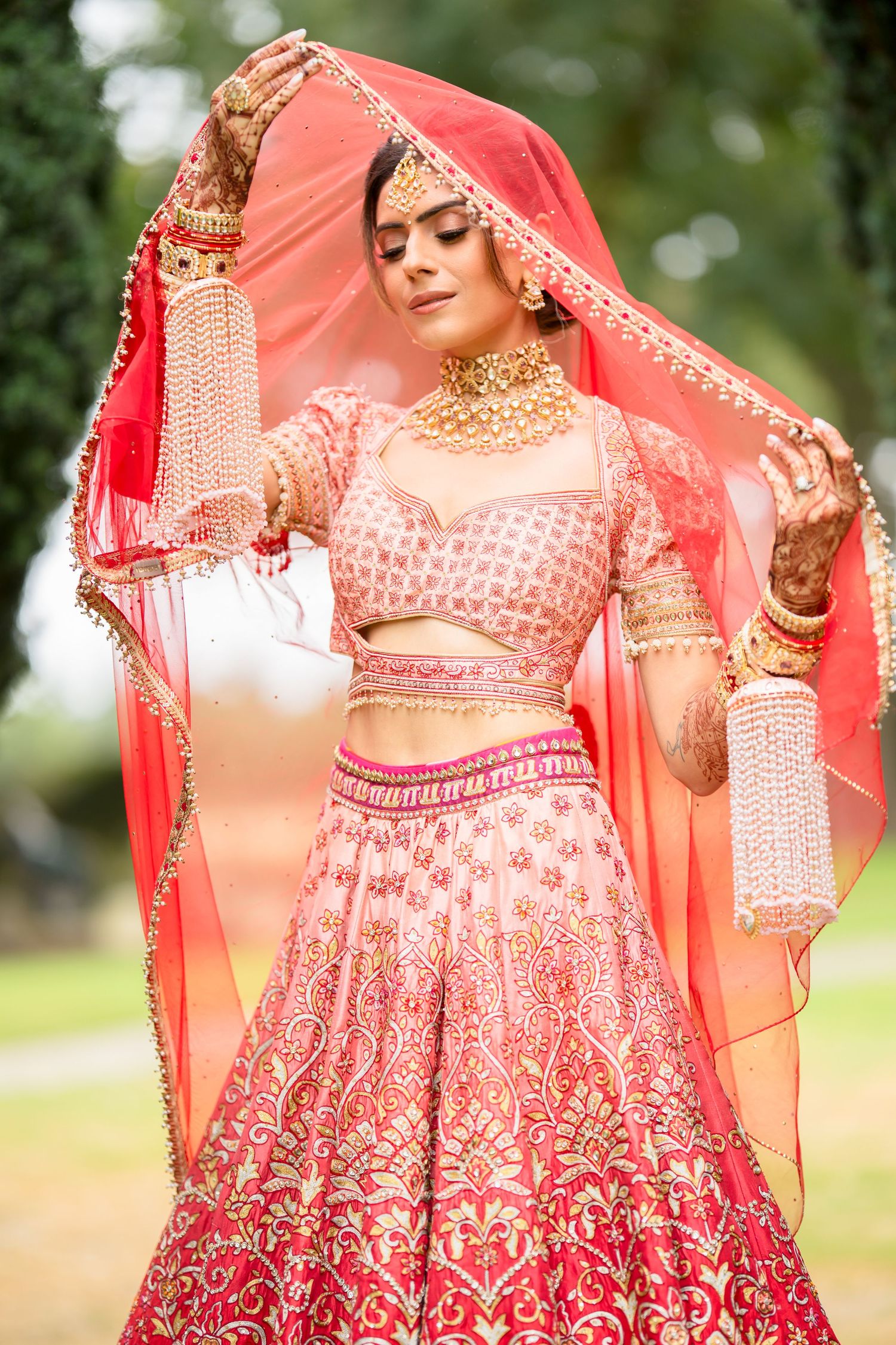 Indian bride in red and pink lehenga with gold embroidery lifts her red dupatta gracefully outdoors.