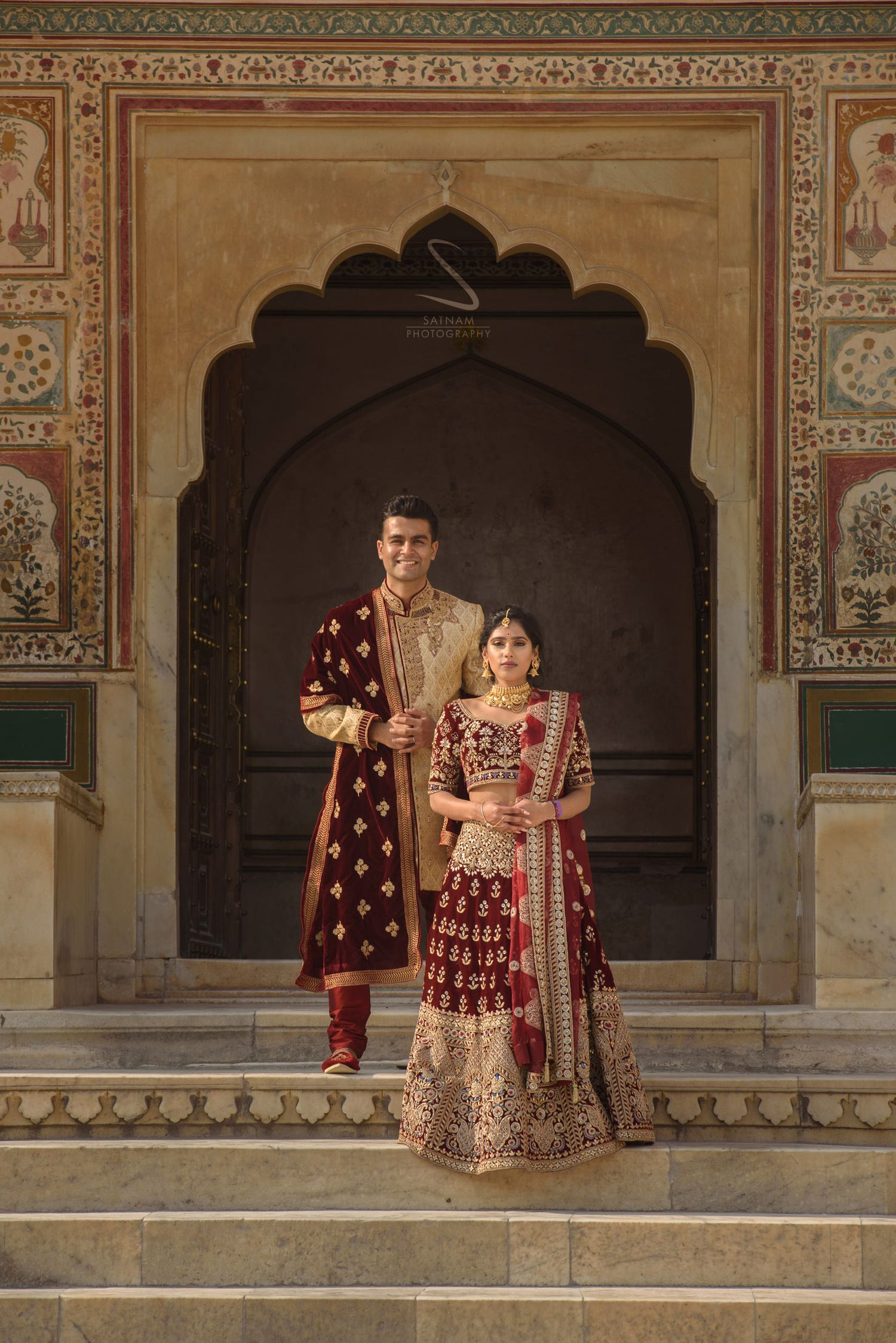 Indian bride and groom in deep maroon bridal outfits posing under ornate Mughal arch doorway.