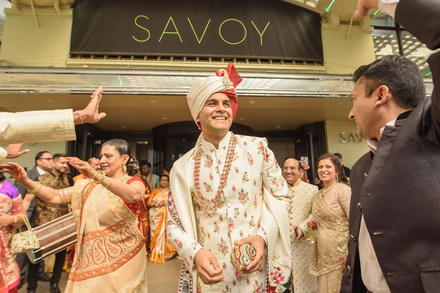 Indian groom in floral sherwani celebrates outside Savoy hotel during traditional wedding baraat ceremony.