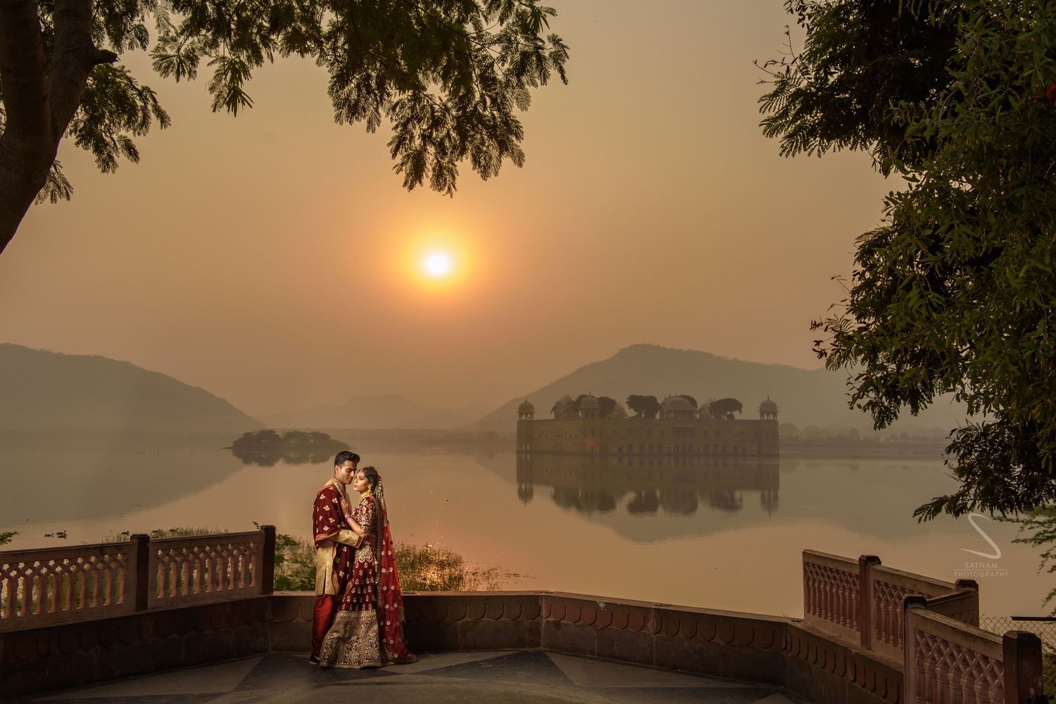 Couple embracing at sunrise by serene Jal Mahal lake with misty hills in Jaipur.