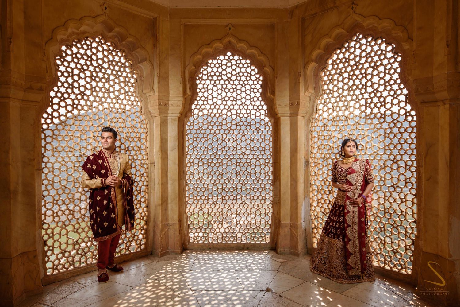 Indian bride and groom in maroon wedding attire posing in stunning latticed stone archways in Jaipur.