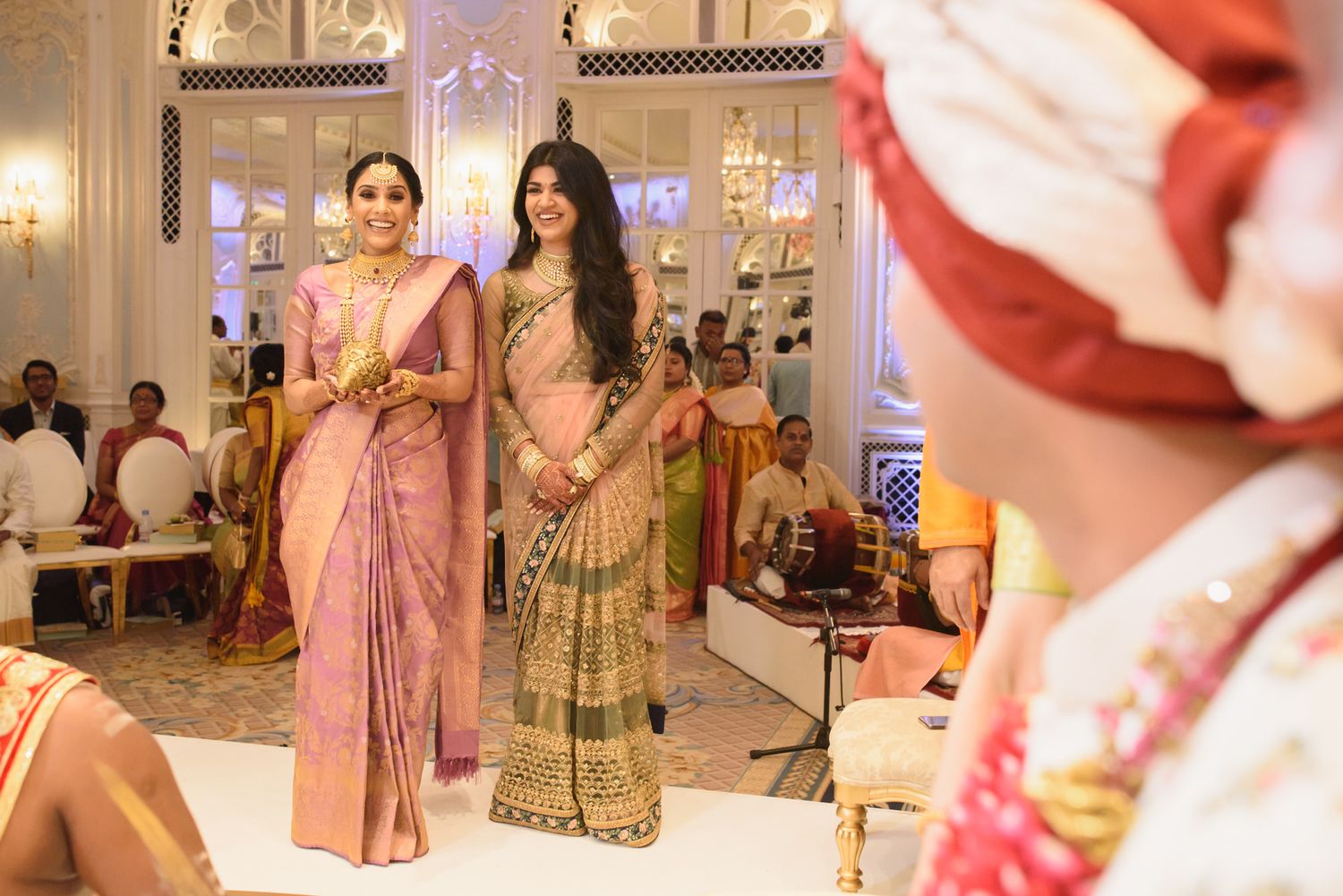 Two elegantly dressed Indian women in sarees smile during a traditional Hindu wedding celebration.