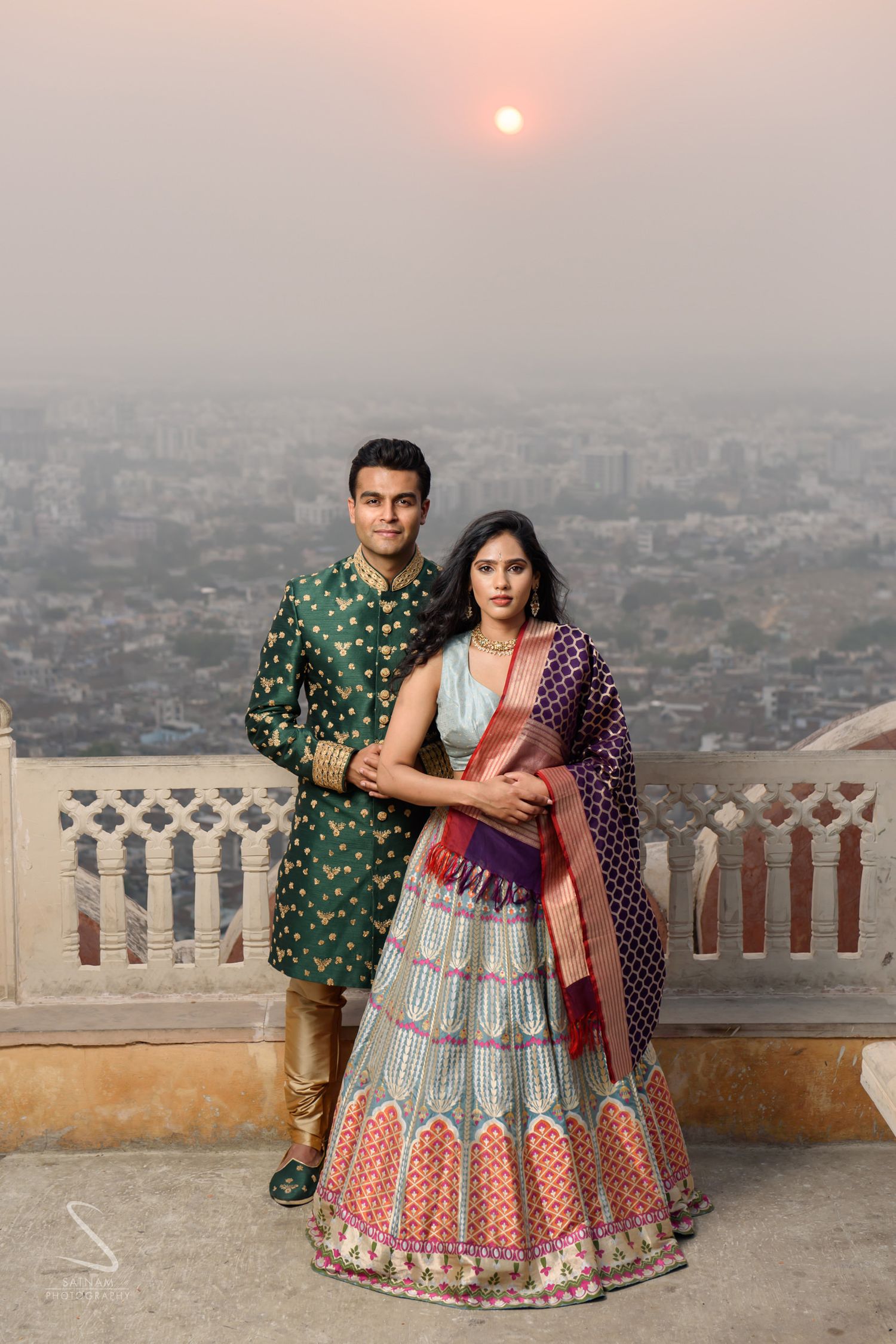 Indian couple in traditional wedding attire posing at sunset with Jaipur city panorama in background.