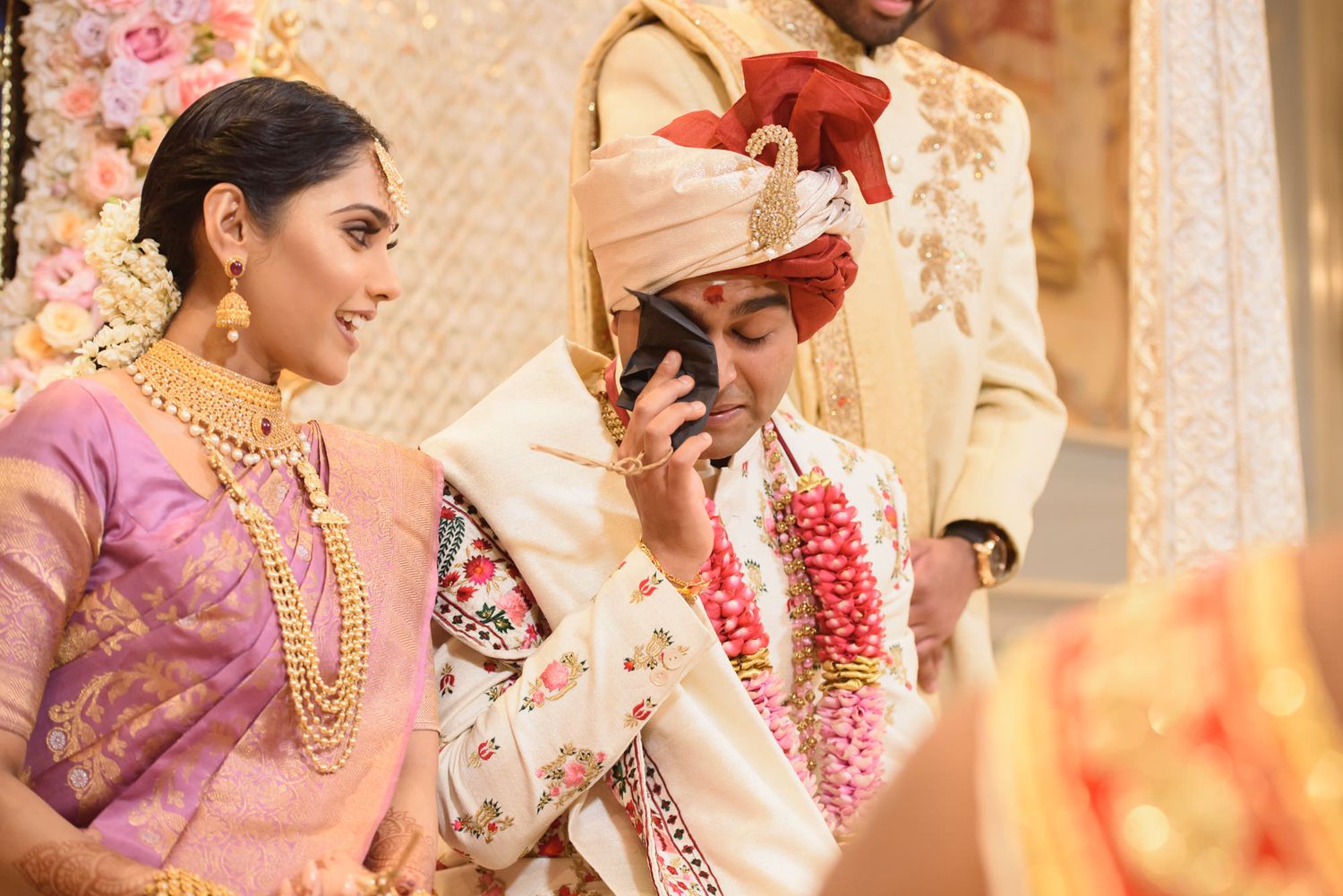 Indian bride in purple saree and groom in red turban share a moment during their Hindu wedding.