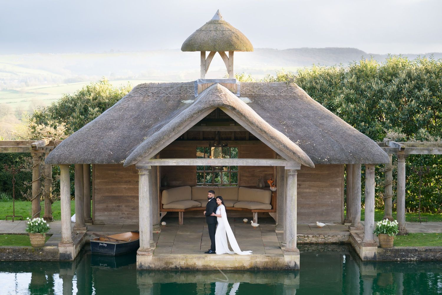 Bride and groom embracing under thatched roof boathouse reflected in calm water with scenic countryside views.