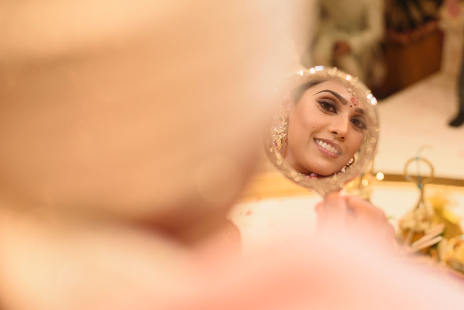 Indian bride in bridal jewelry smiles at her reflection in handheld mirror.