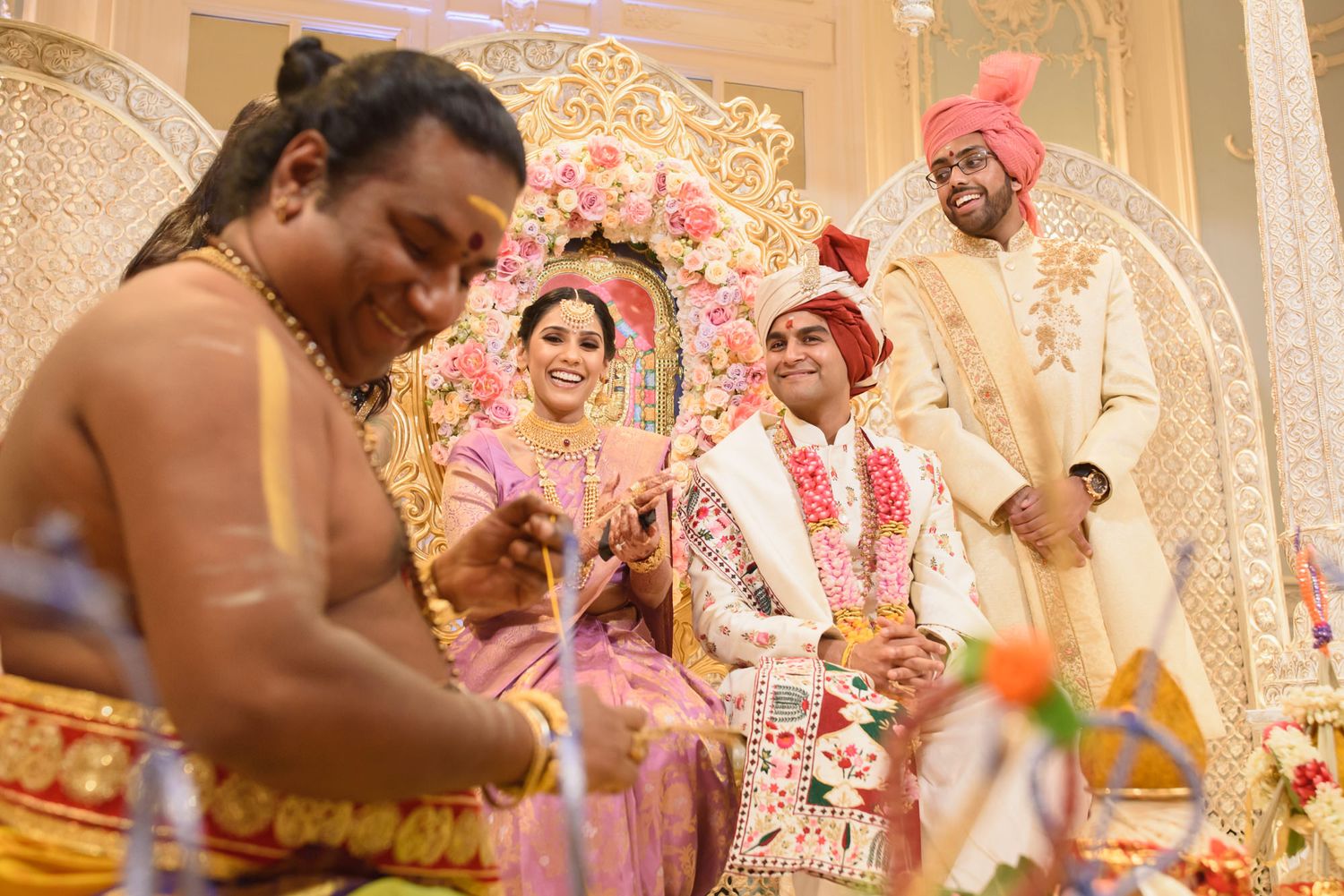 Hindu priest performs wedding rituals as smiling bride and groom celebrate during traditional Indian ceremony.