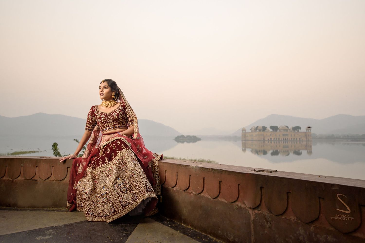 Indian bride in stunning maroon and gold lehenga sitting by Jal Mahal lake palace at dusk in Jaipur.