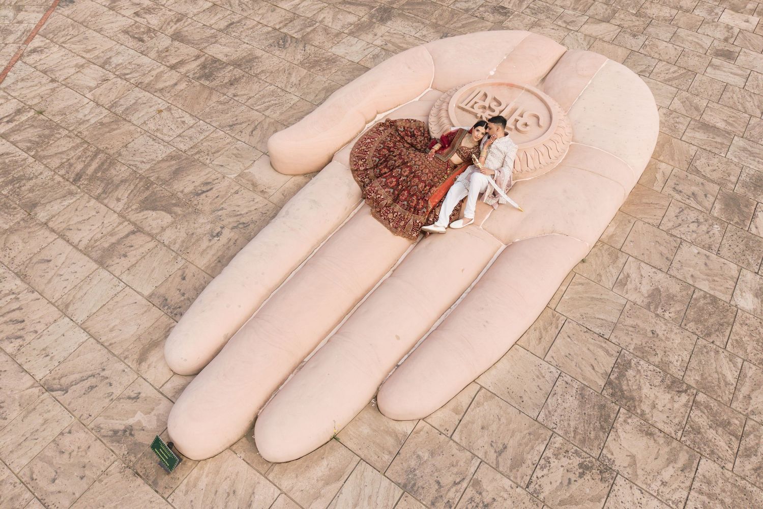 Large pink hand sculpture on a tiled plaza holding miniature figurines in traditional attire.