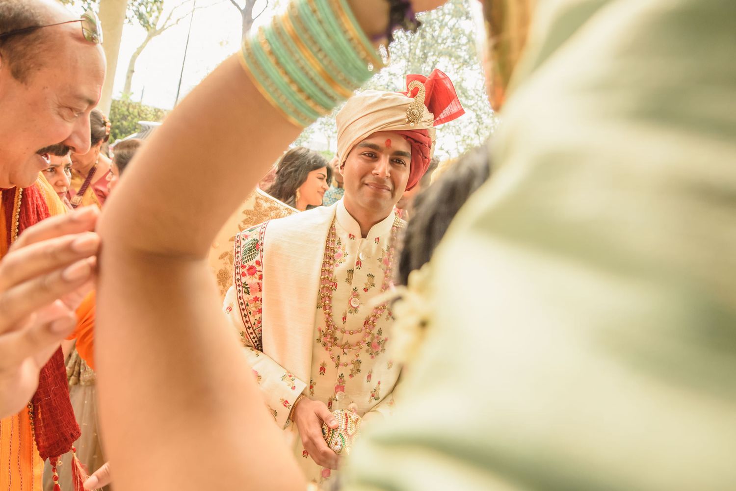 Smiling Indian groom in cream sherwani and red turban during colorful traditional wedding baraat.