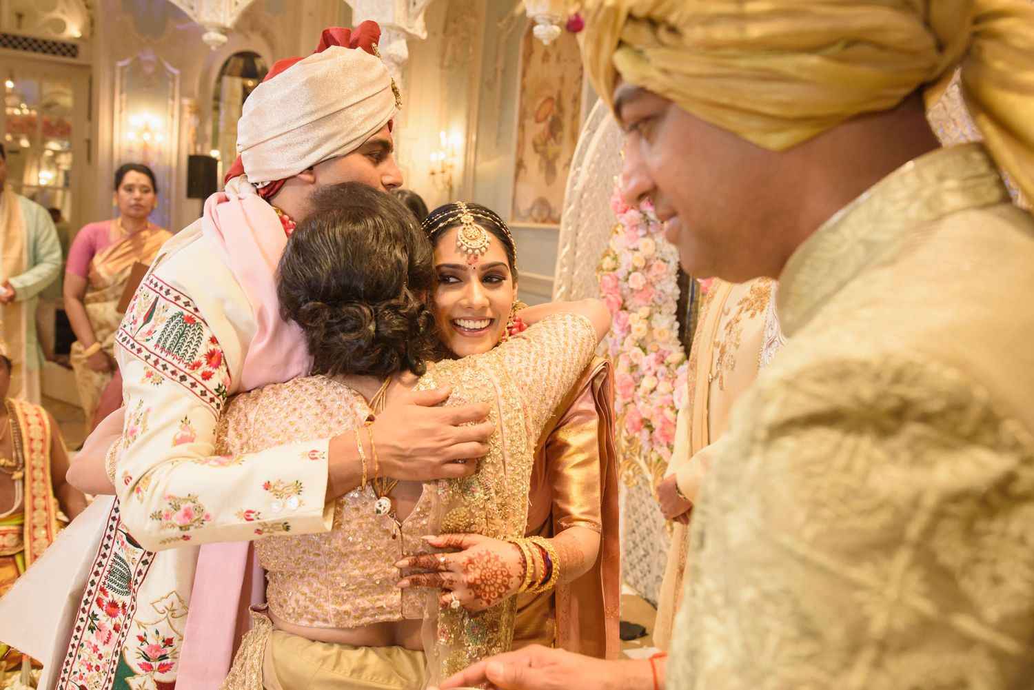 Bride in gold lehenga embracing guests at traditional Indian wedding ceremony with floral decorations.