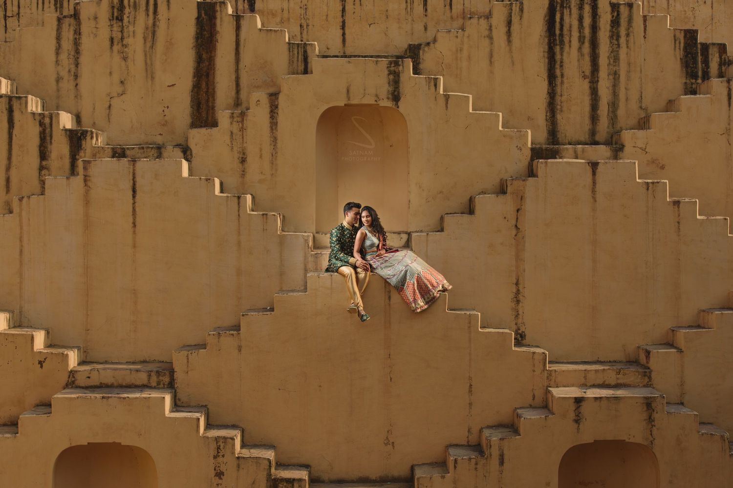 Couple in colorful Indian outfits sitting together on the geometric steps of Panna Meena Ka Kund stepwell.