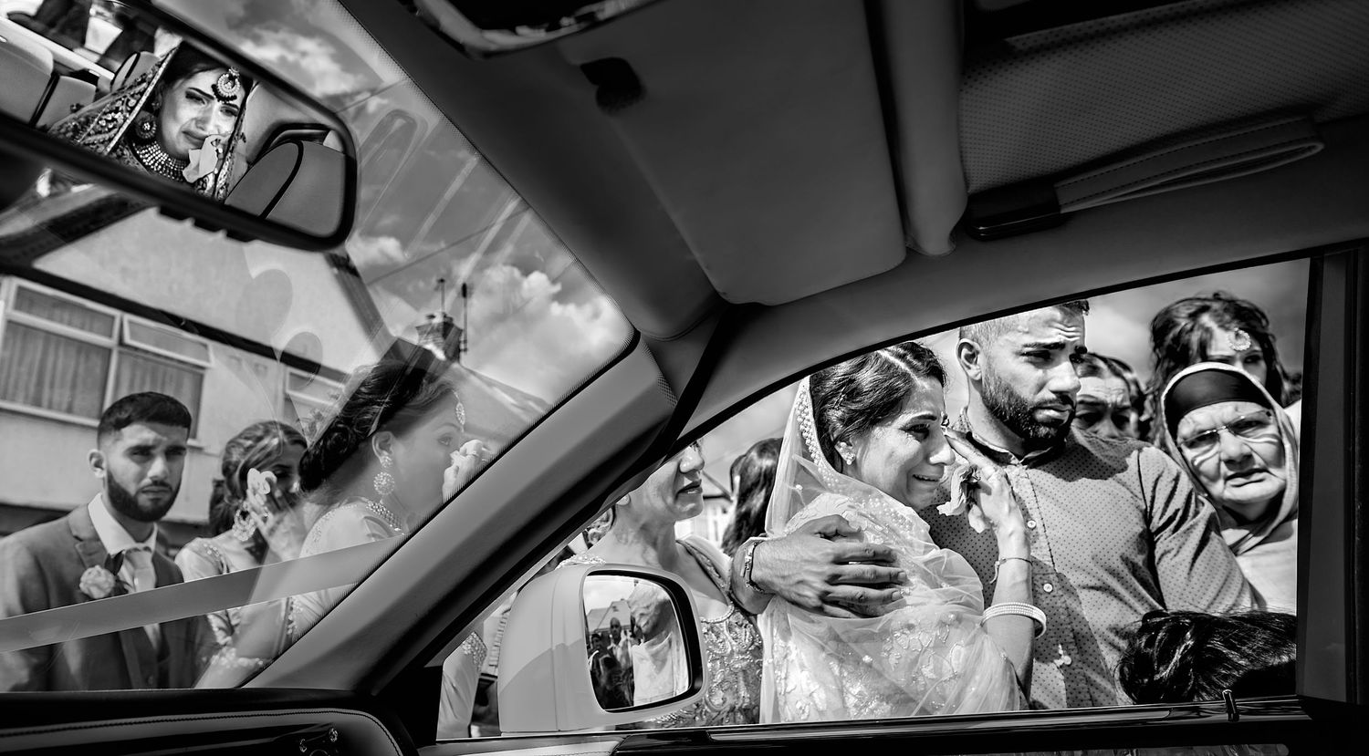 A black and white photo captures an emotional bride embracing family members through a car window.