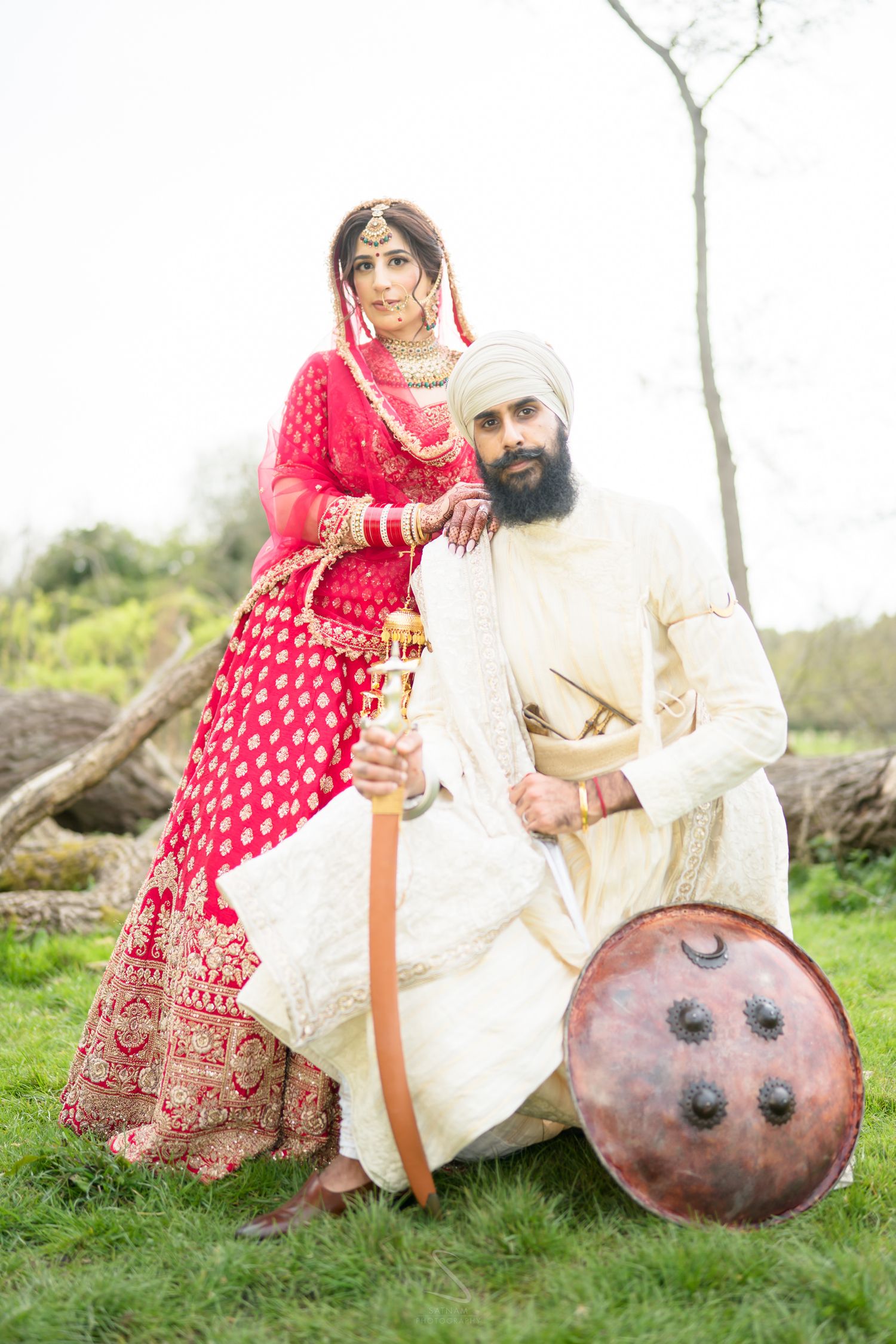Sikh bride in red lehenga and groom in white sherwani posing with traditional sword and shield outdoors.