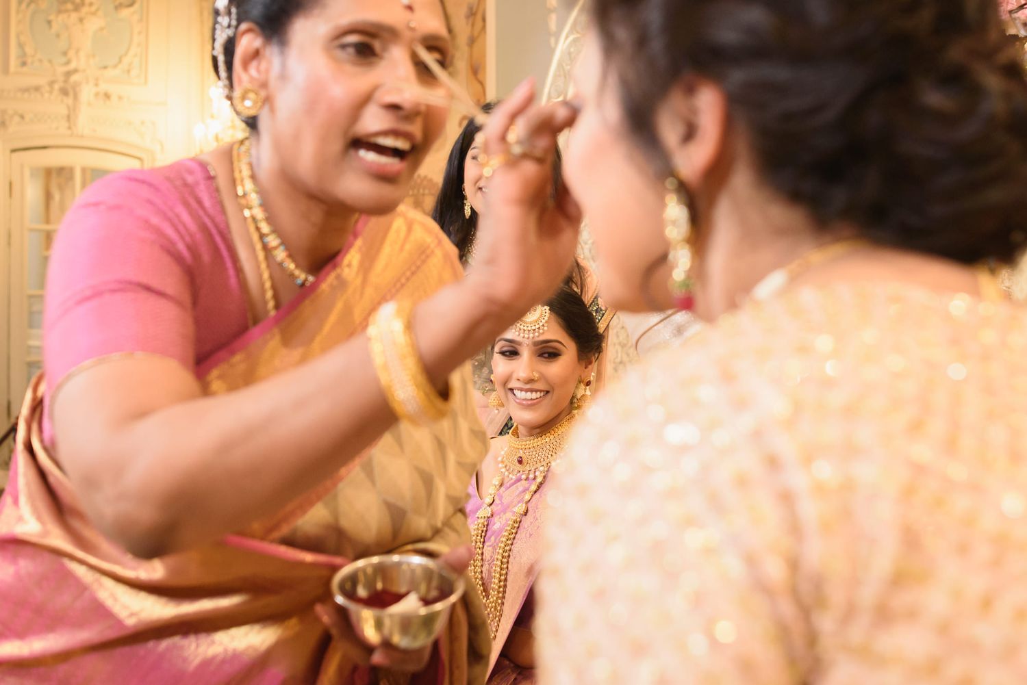 Woman in pink saree applies sindoor to bride during traditional South Indian wedding ritual ceremony.