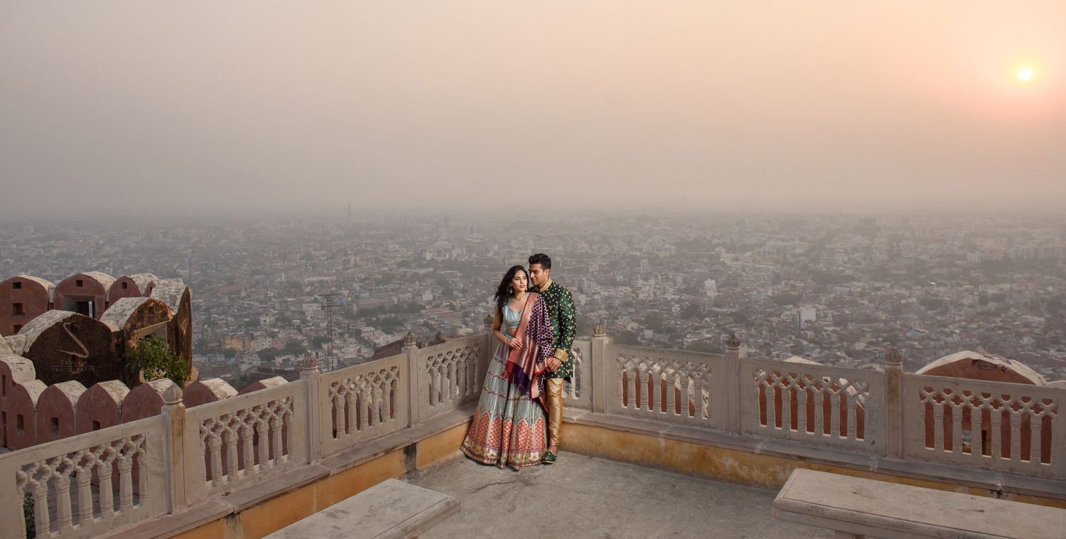 Couple in vibrant Indian wedding outfits embracing on a rooftop terrace overlooking Jaipur city at sunset.