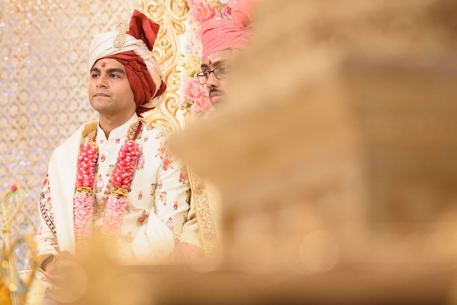 Indian groom in red and gold turban with floral garland during traditional Hindu wedding ceremony.