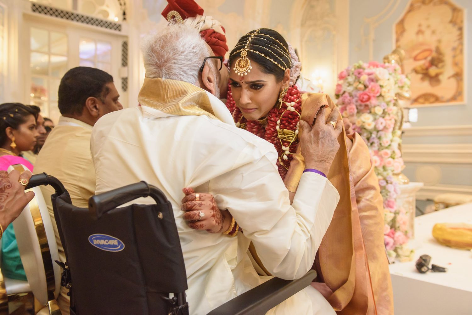 Bride in red bridal jewelry tenderly embracing elderly man in wheelchair at Indian wedding ceremony.