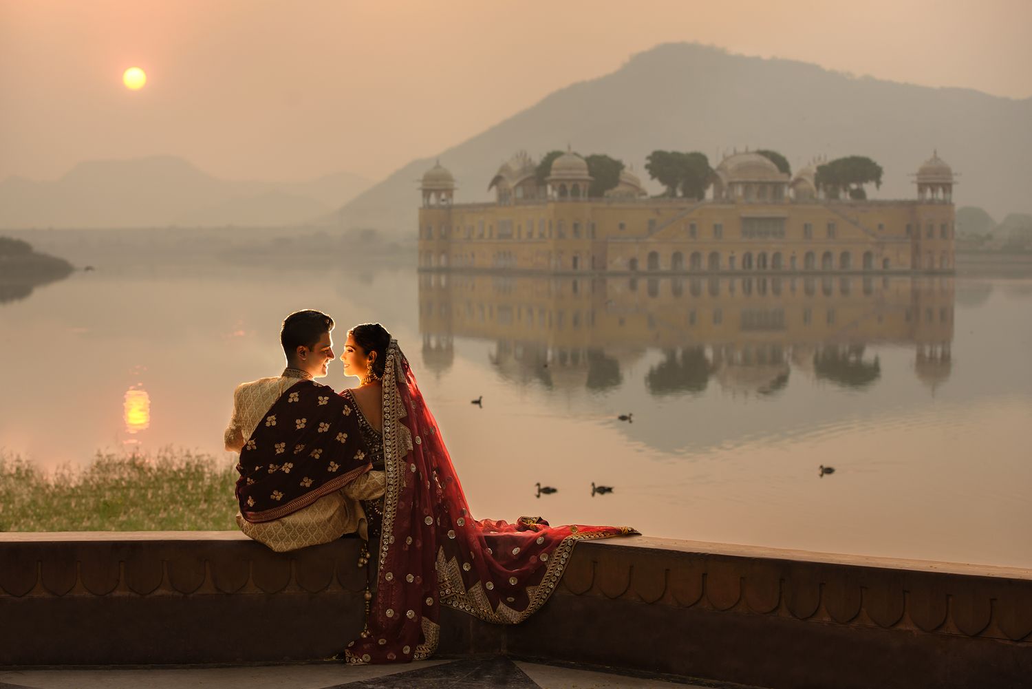 Indian couple in traditional attire sit by lake at sunset near Jal Mahal Jaipur.