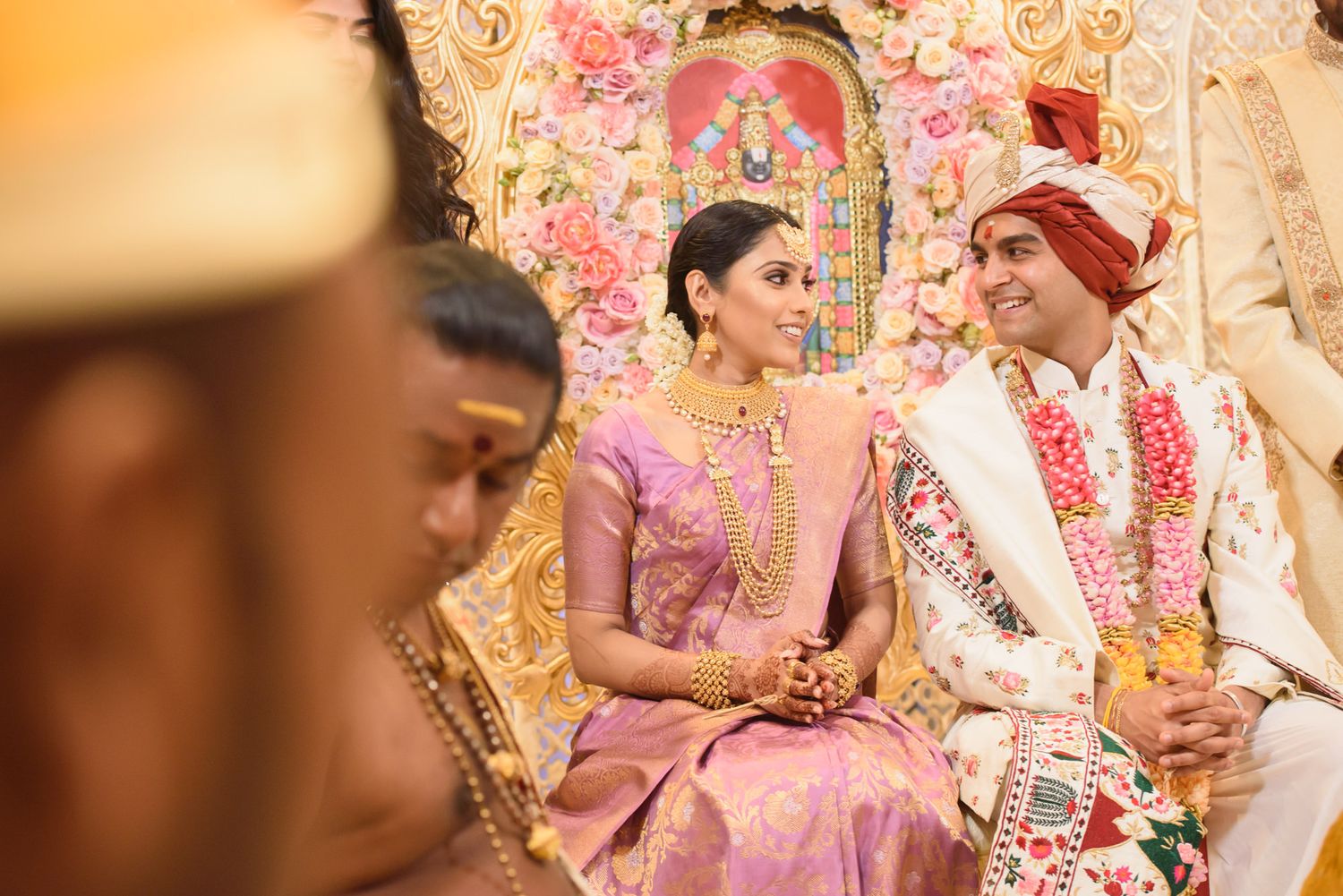 Bride in purple silk saree and groom in white sherwani sit together during traditional Hindu wedding ceremony.