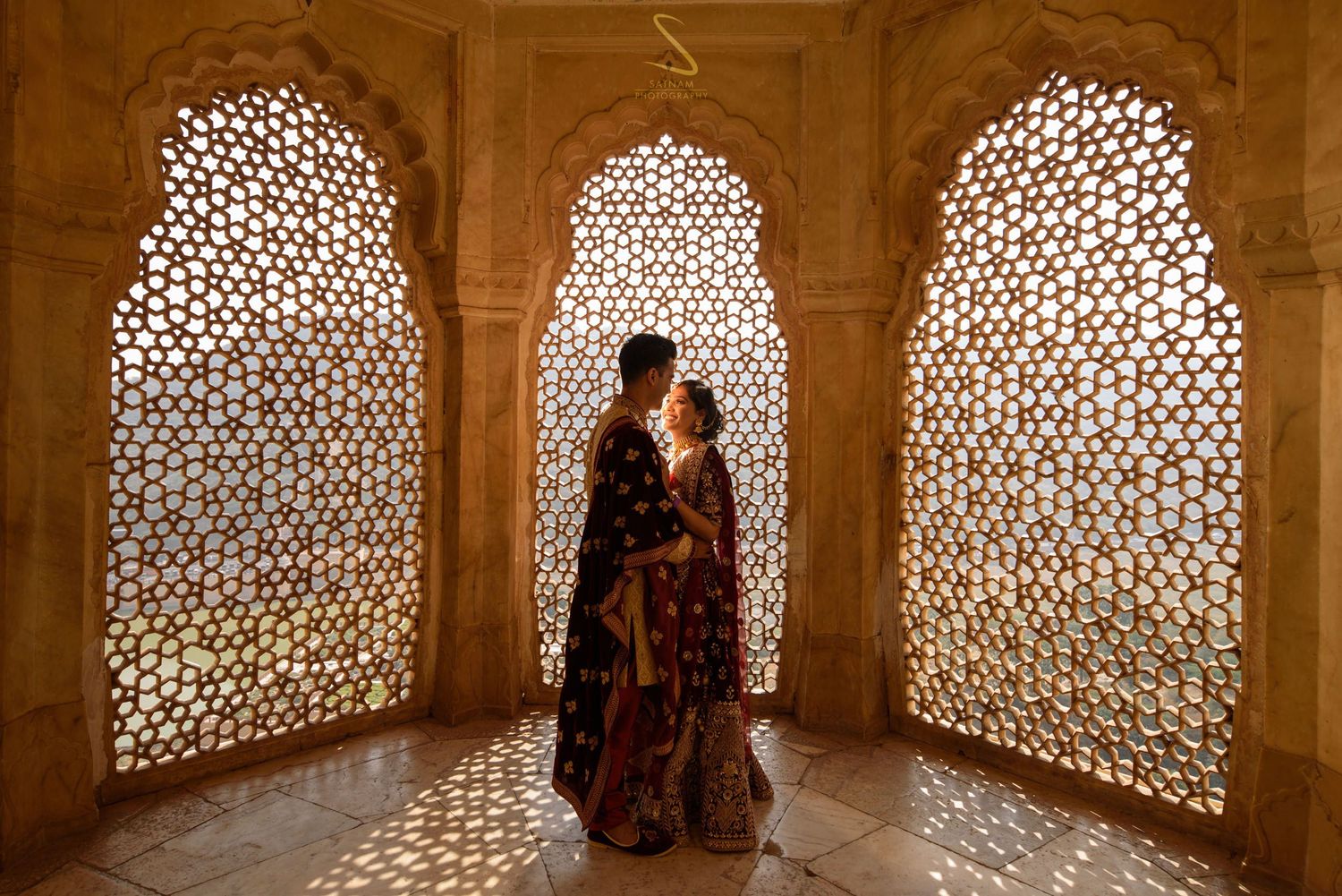 Couple silhouetted against ornate latticed stone windows in a golden Rajasthani palace interior.