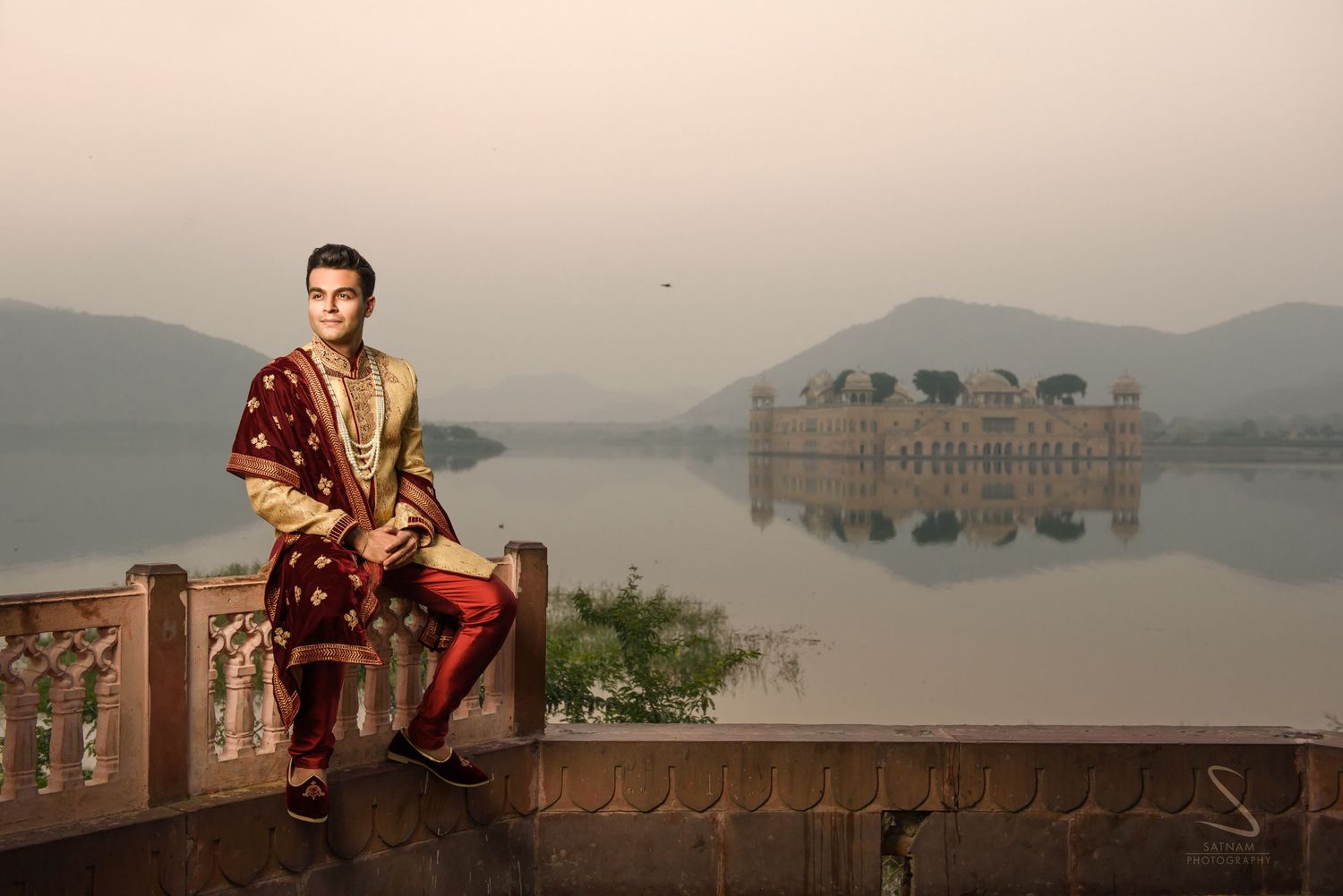 Man in traditional Indian sherwani sitting by serene lake with Jal Mahal palace backdrop.