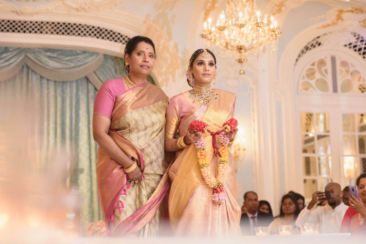 Bride and older woman in silk sarees at elegant Indian wedding reception hall.