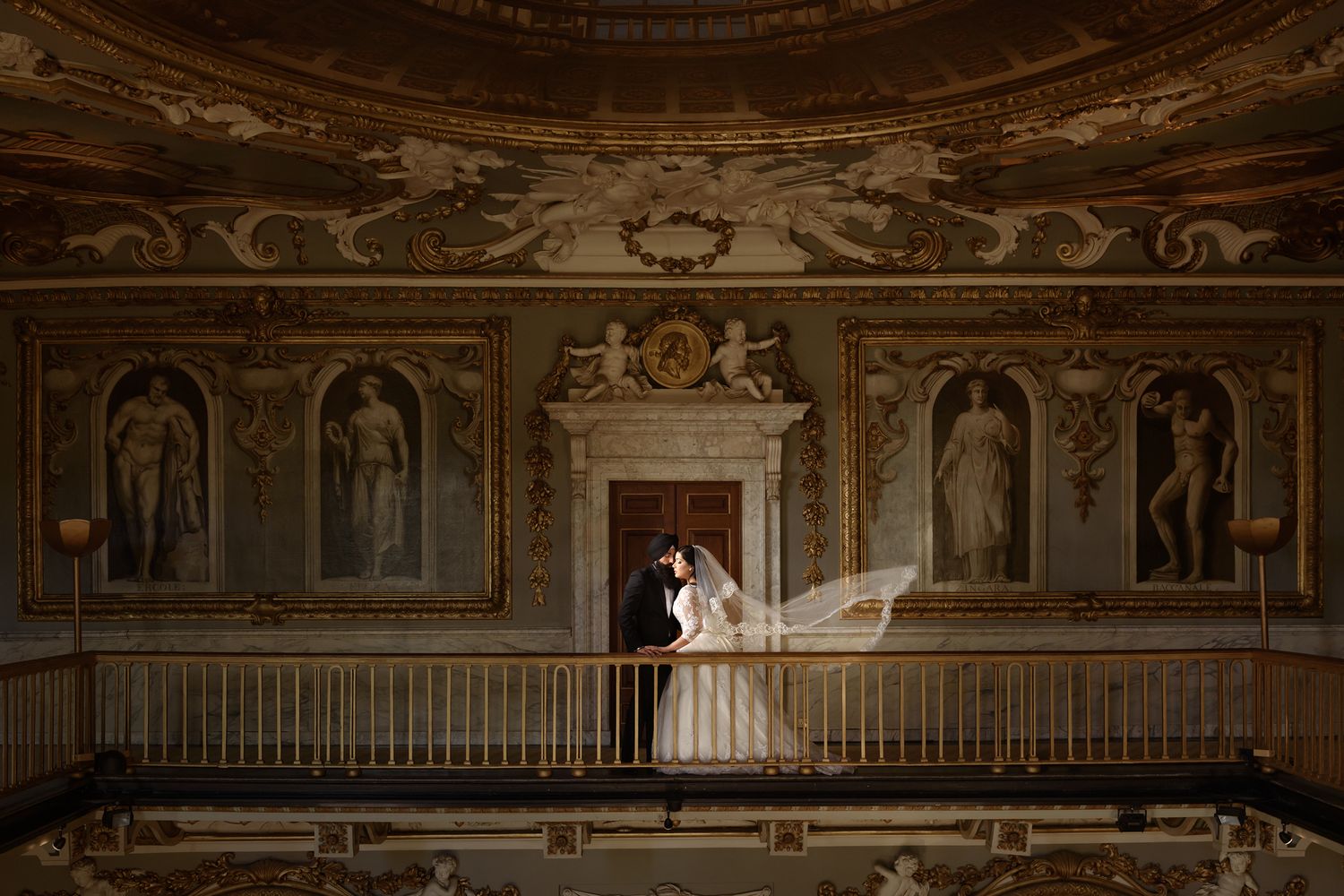 Bride and groom share romantic moment on ornate balcony in grand historic venue with elaborate ceiling murals.