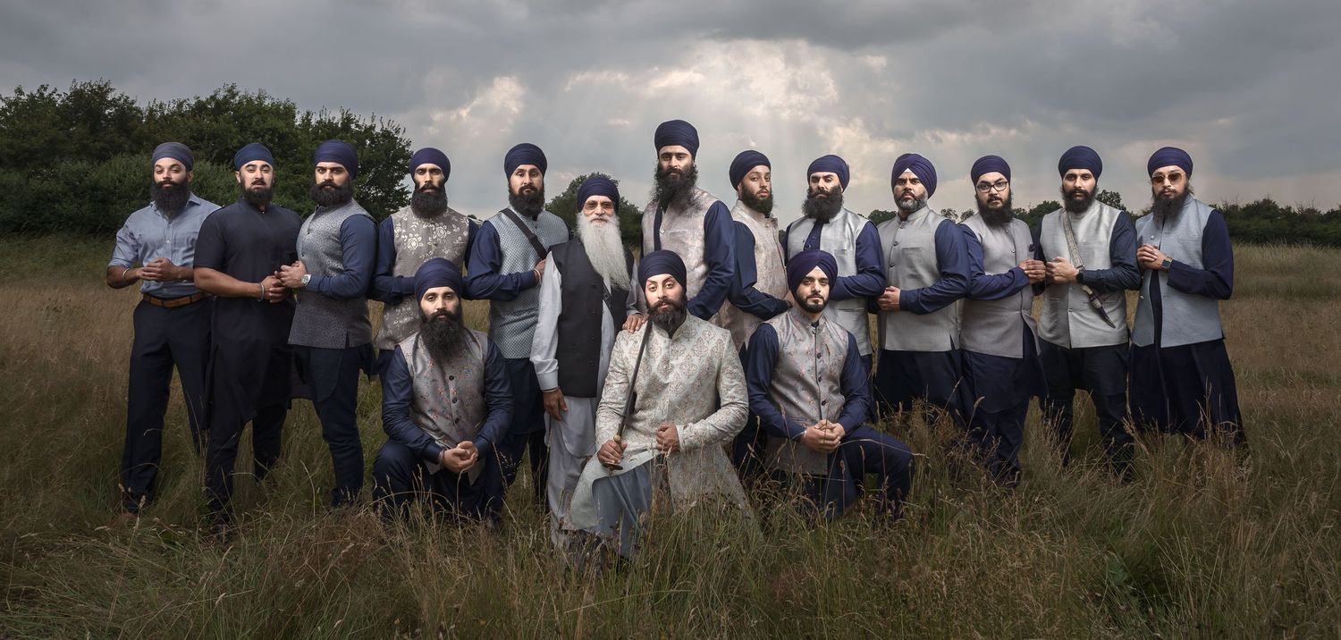 A group of Sikh groomsmen with beards and turbans pose together in a field under a dramatic cloudy sky.