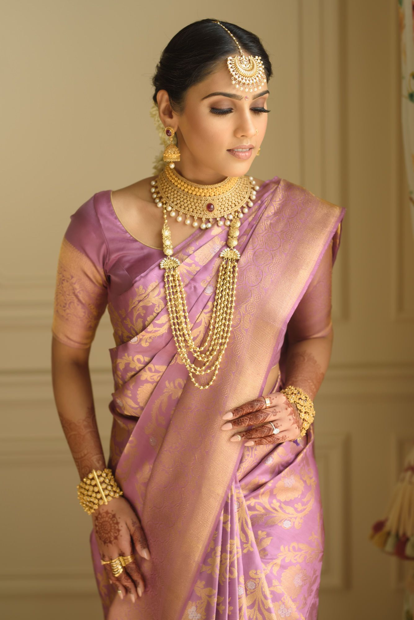 South Indian bride in lavender Kanjivaram saree with layered gold necklaces and maang tikka.