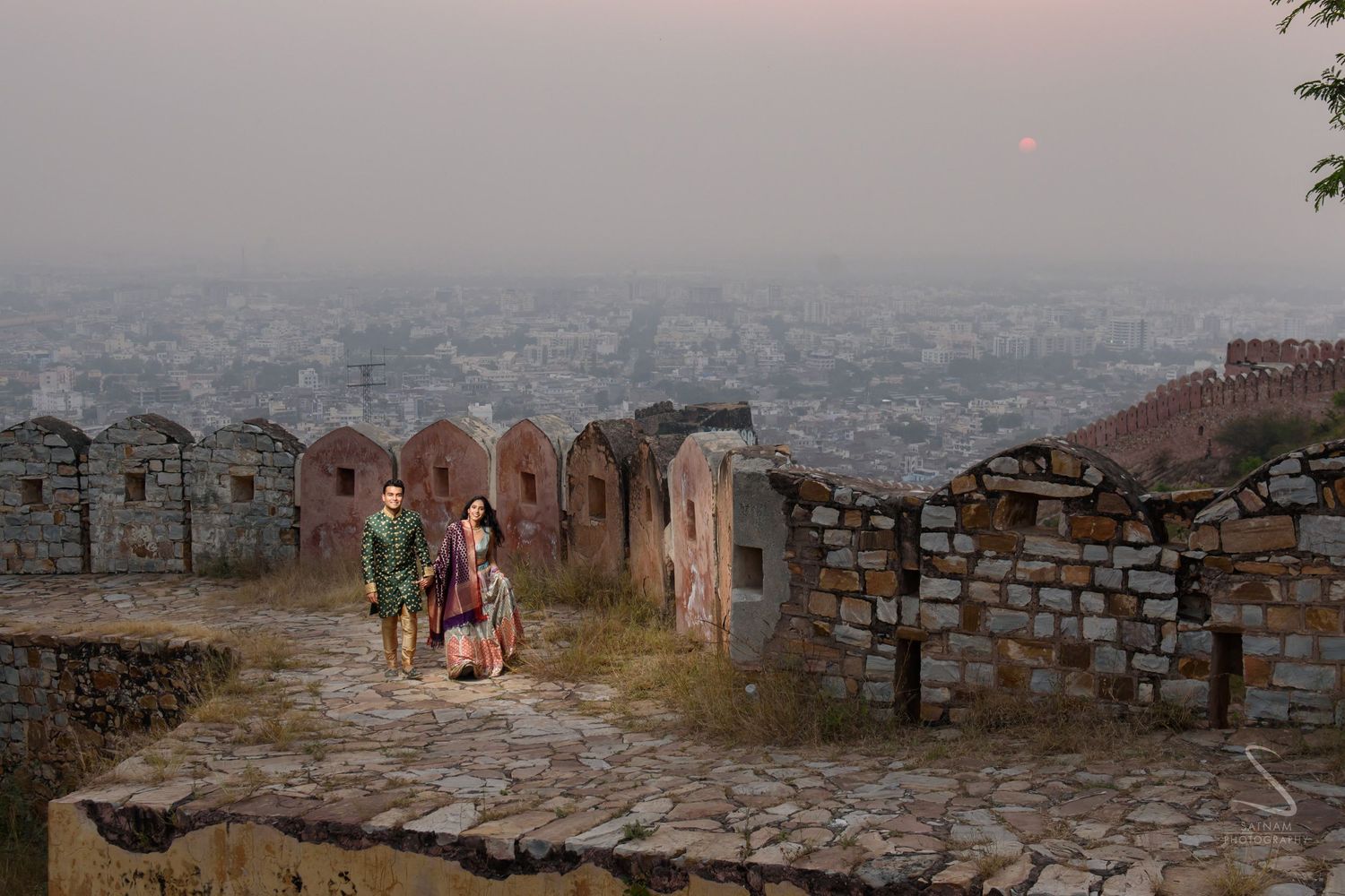 Couple in traditional Indian wedding clothes walking along historic fort walls at dusk in Jaipur.